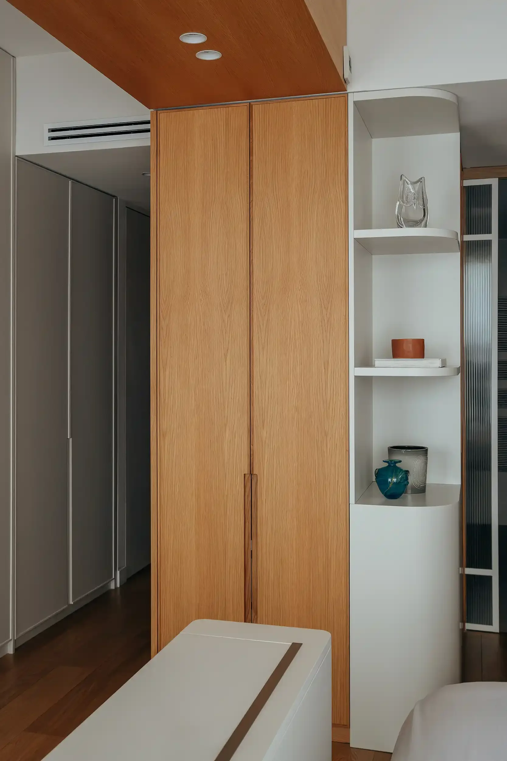 Armoire de rangement en chêne clair jouxtant une bibliothèque blanche aux étagères arrondies avec objets de décoration.