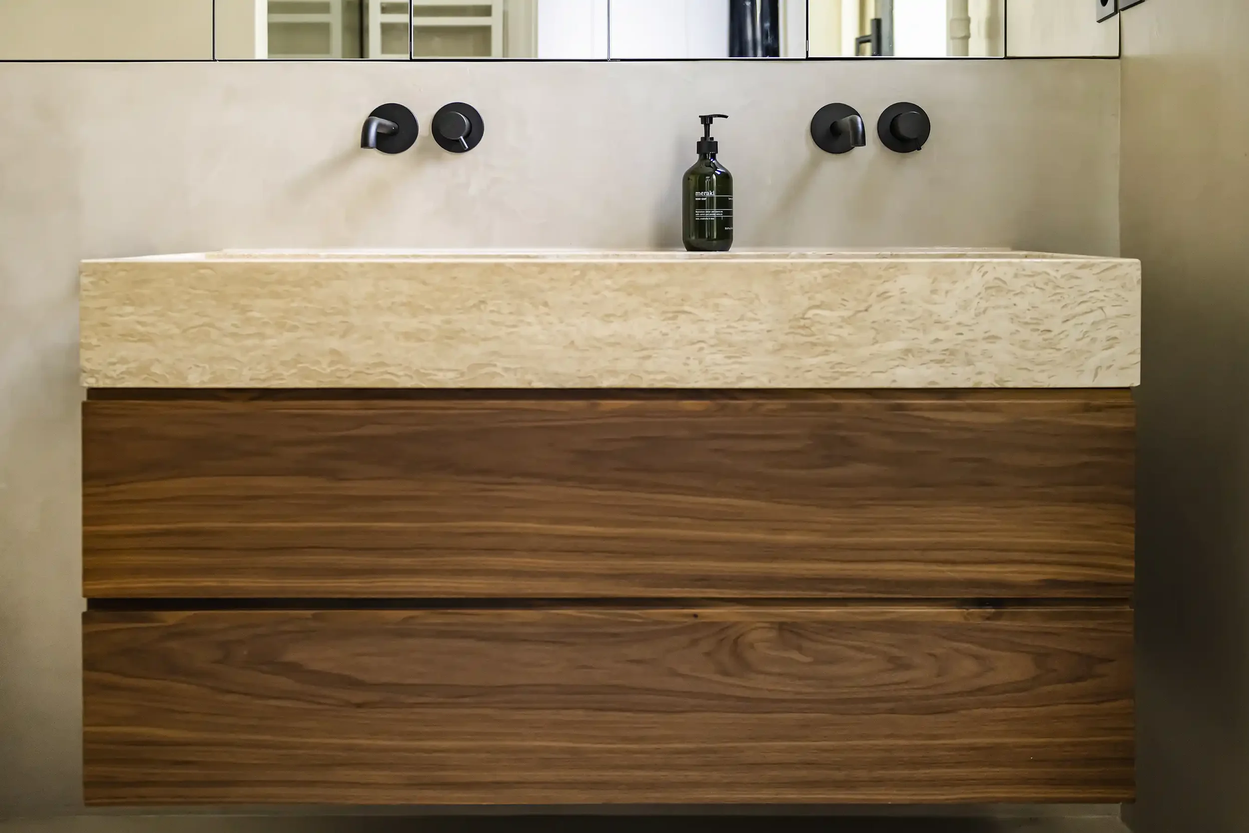 Détail d'une salle de bain moderne avec une vasque en pierre naturelle beige posée sur un meuble suspendu en bois de noyer avec robinetterie noire encastrée.