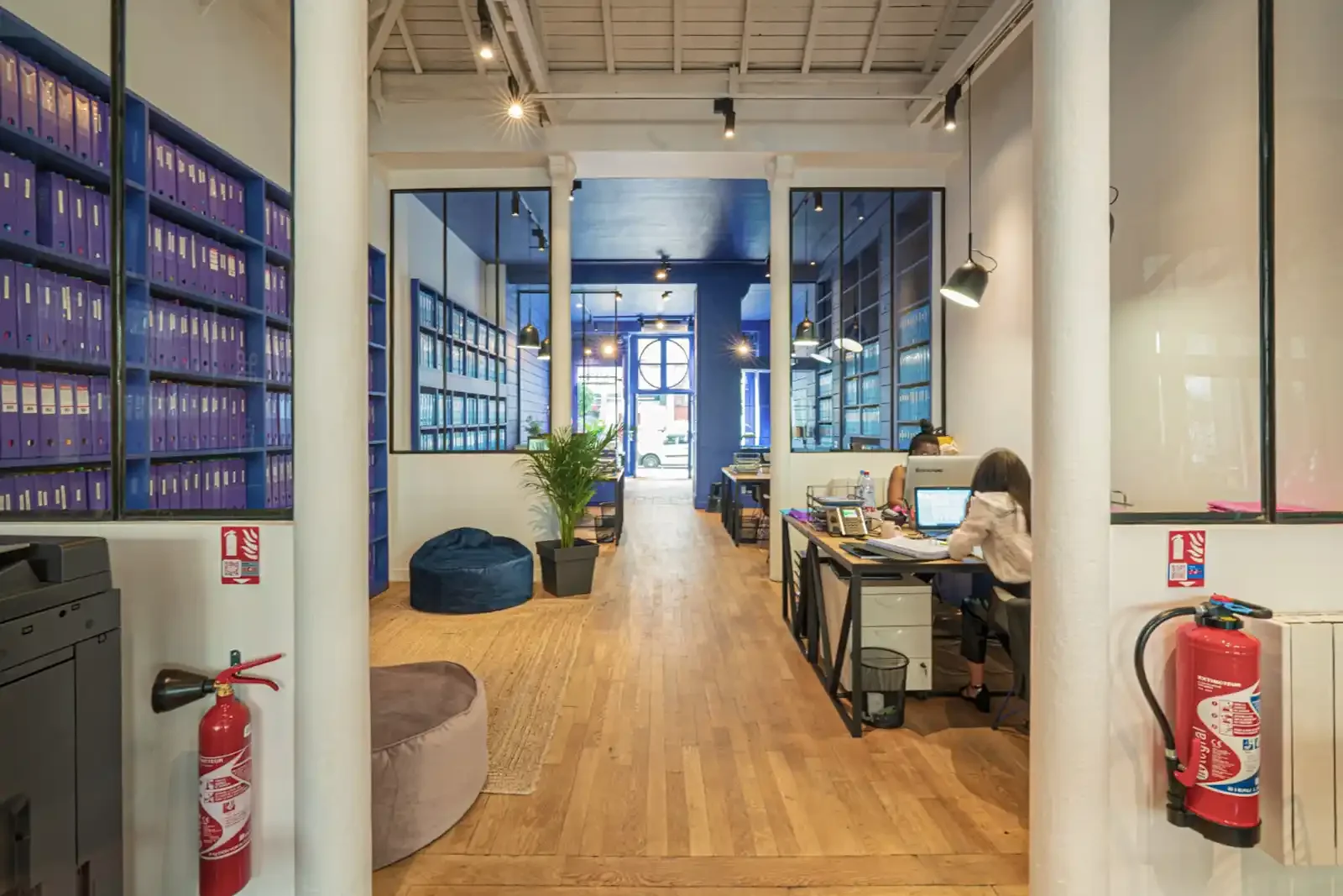 Grand open space lumineux d'une agence immobilière avec parquet en bois, verrières intérieures et colonnes de style industriel.
