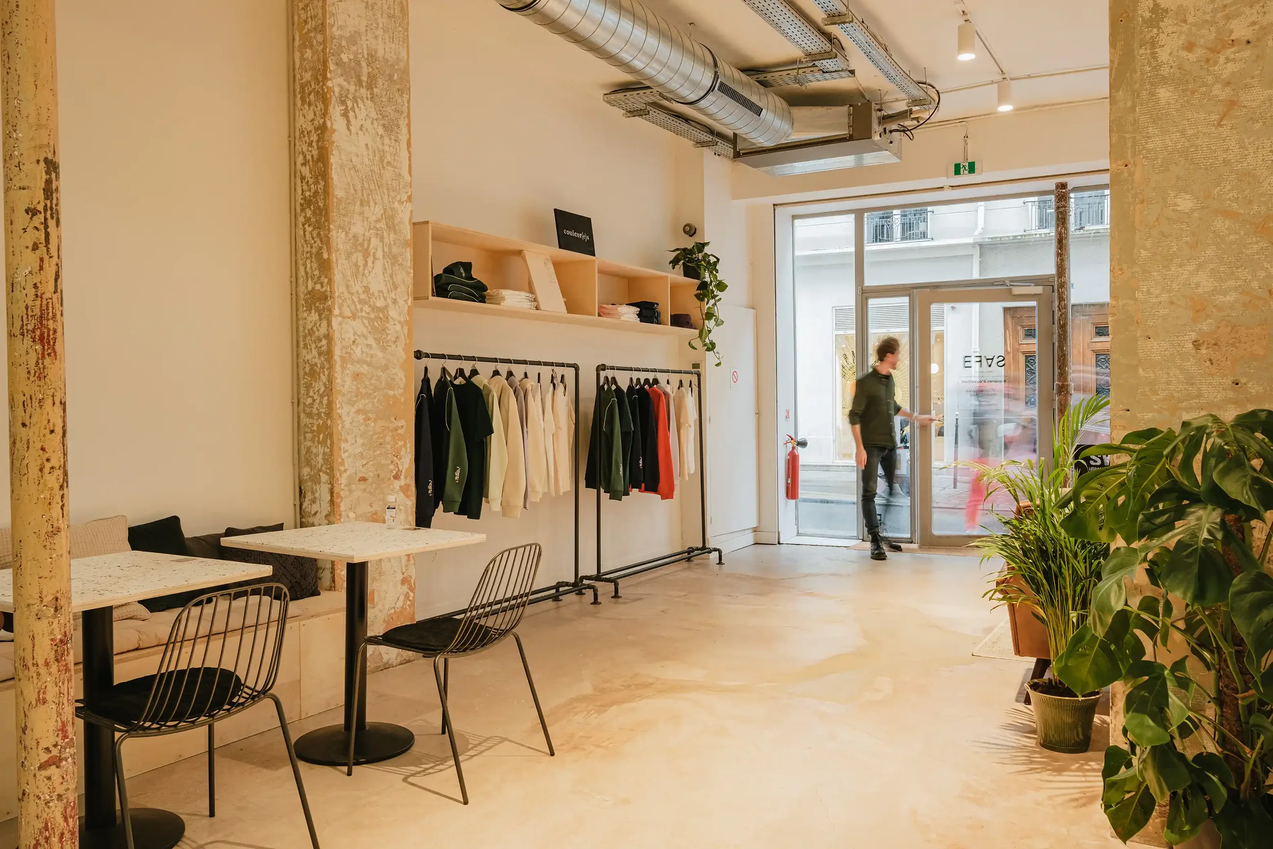 Vue de l'entrée d'un concept store parisien avec grandes baies vitrées, tables de coworking, plantes vertes et portants de vêtements de créateurs.
