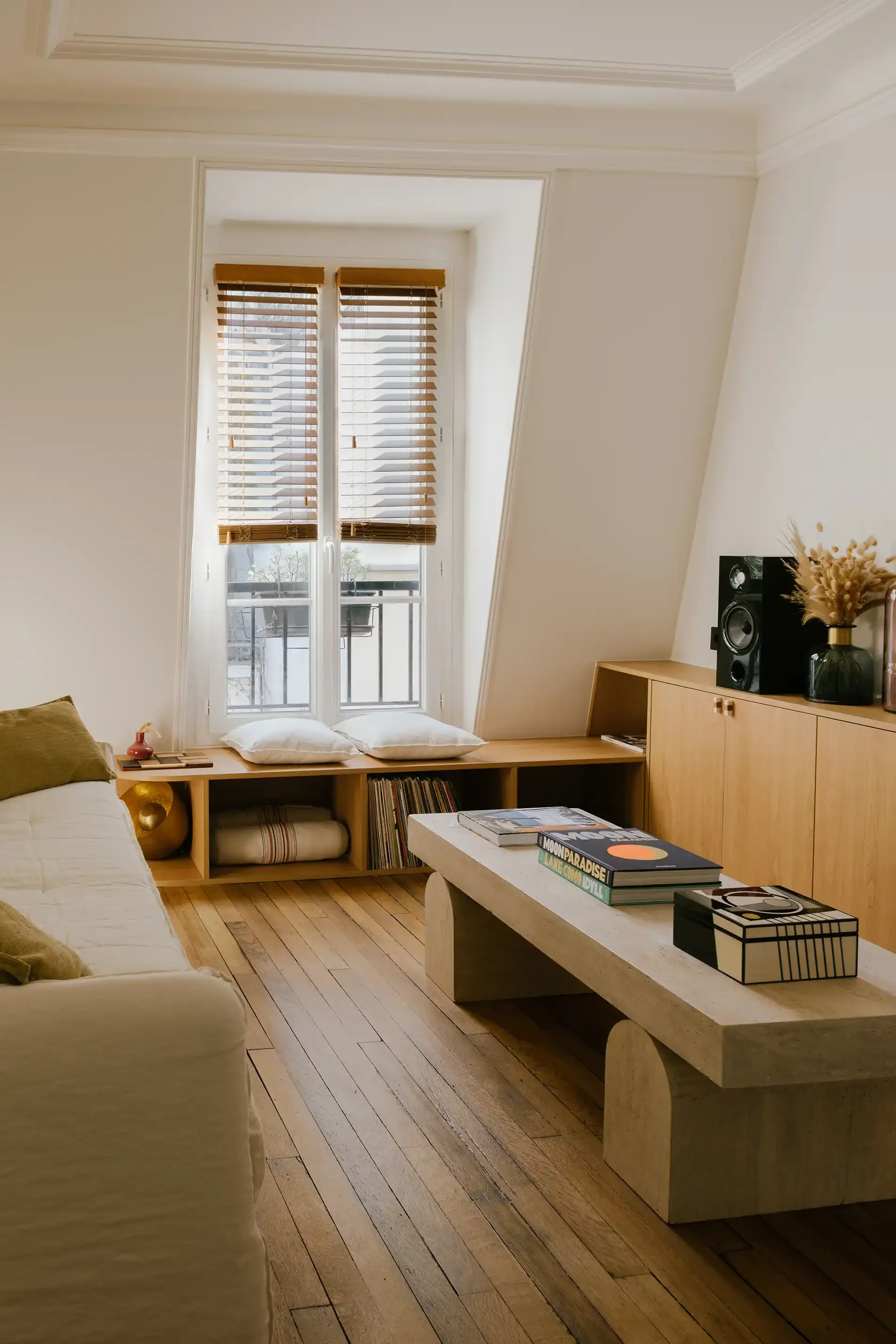 Aménagement d'un salon lumineux avec table basse en travertin, canapé blanc et bibliothèque sur mesure sous une fenêtre mansardée.