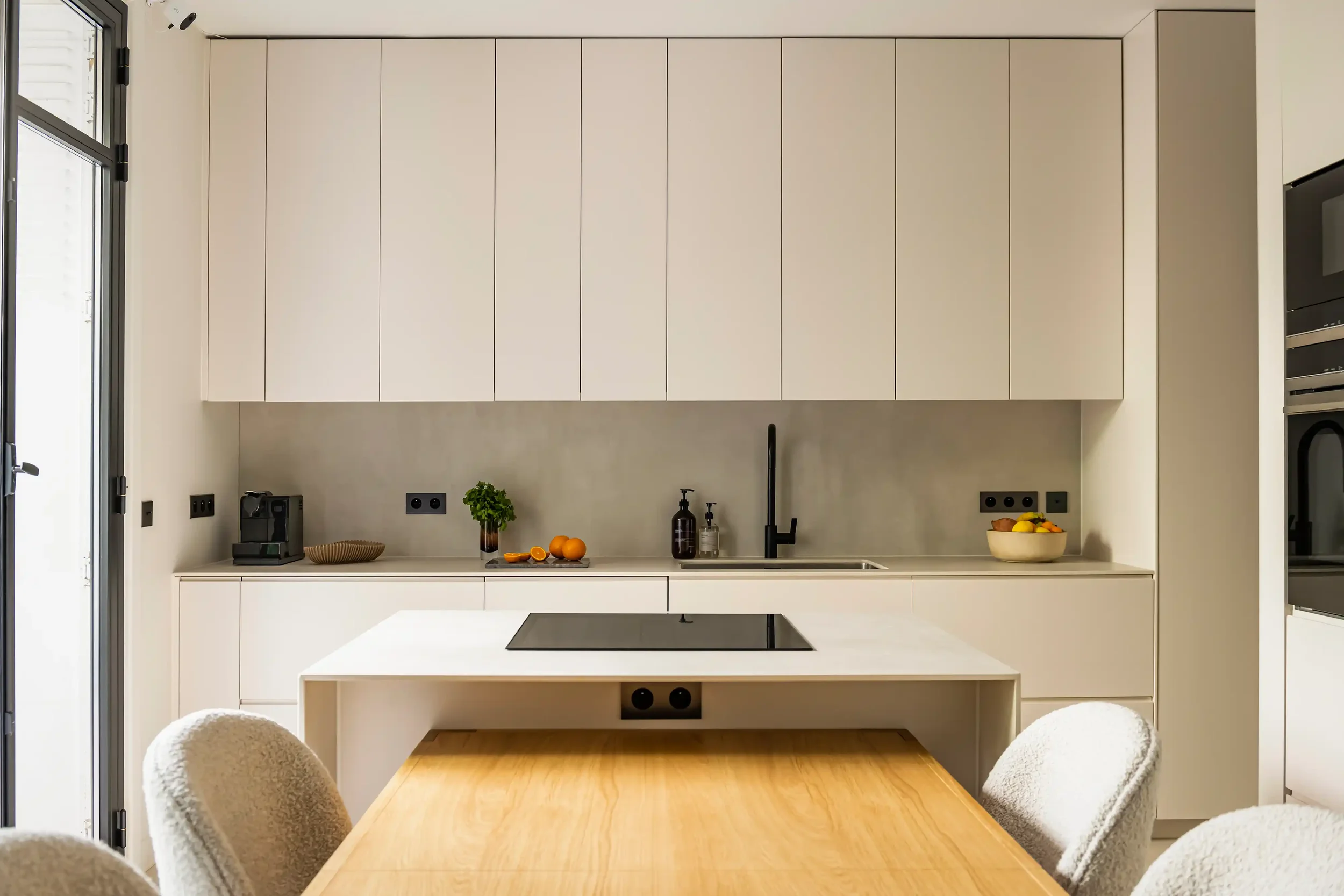 Vue de face d'une cuisine minimaliste avec crédence en pierre beige, robinetterie noire et plaque de cuisson intégrée.