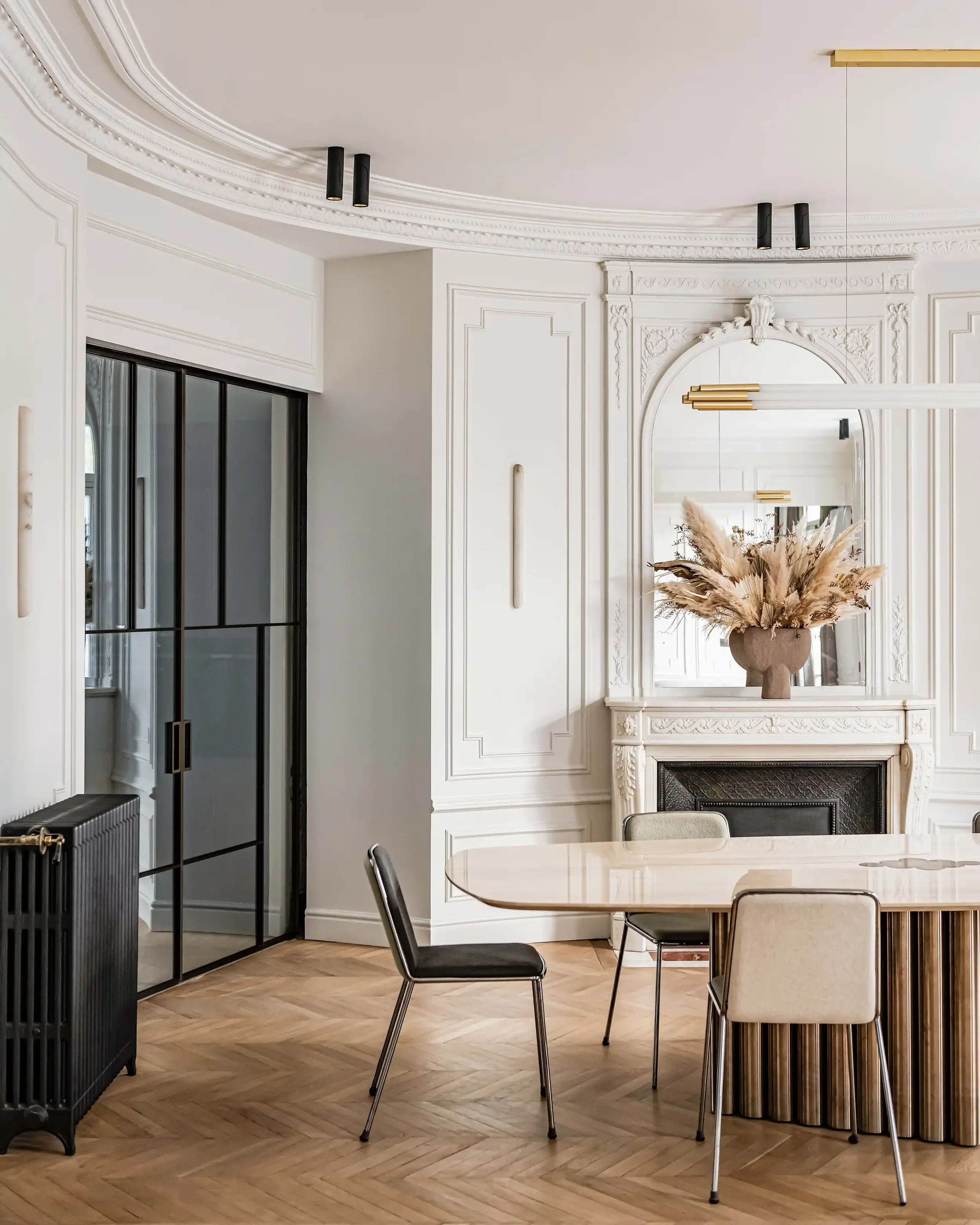 Vue sur une salle à manger ouverte avec cheminée ancienne en marbre, miroir trumeau et verrière style atelier en métal noir.