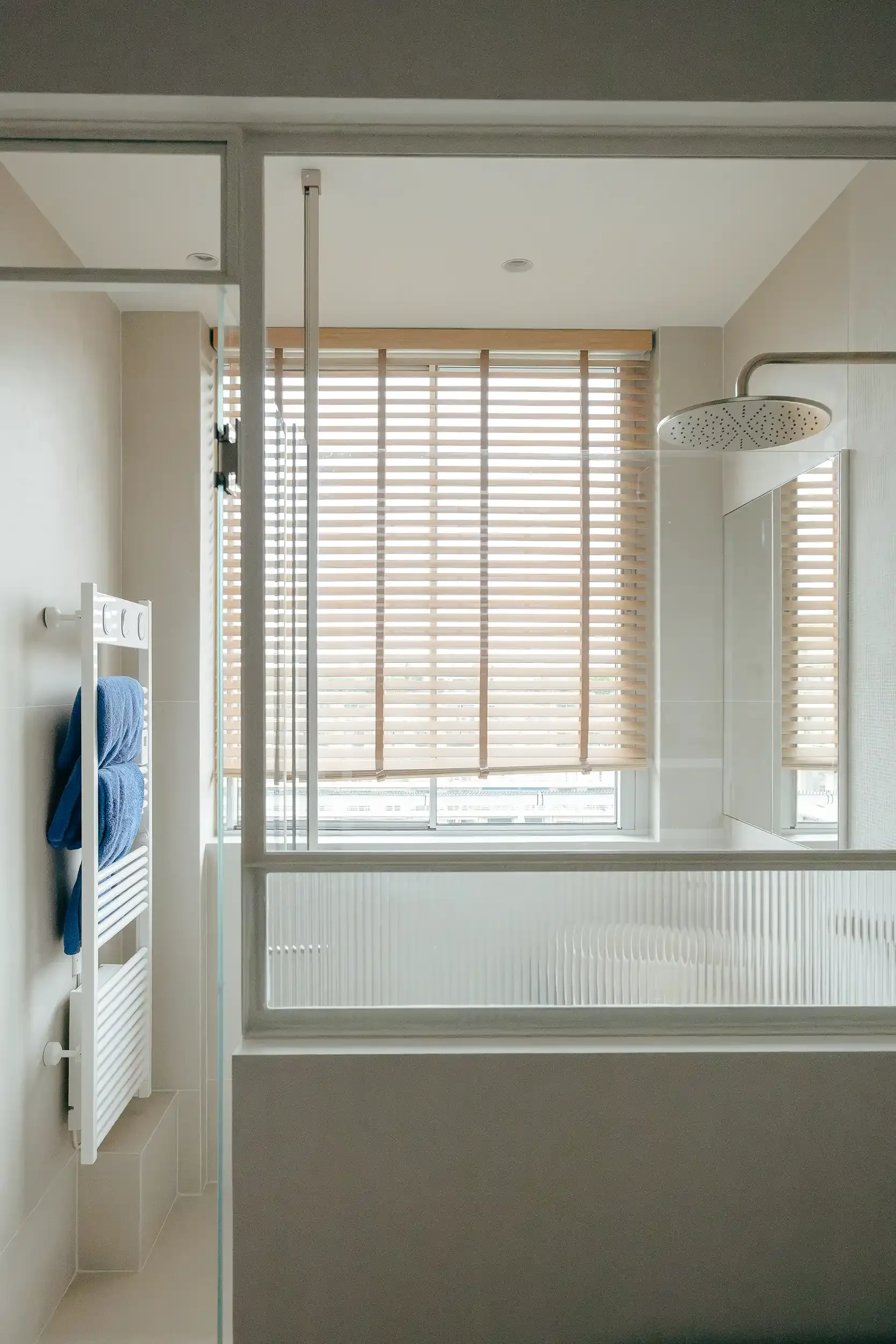 Salle de bain lumineuse avec paroi de douche en verre flûte, store vénitien en bois et ciel de pluie en acier brossé.