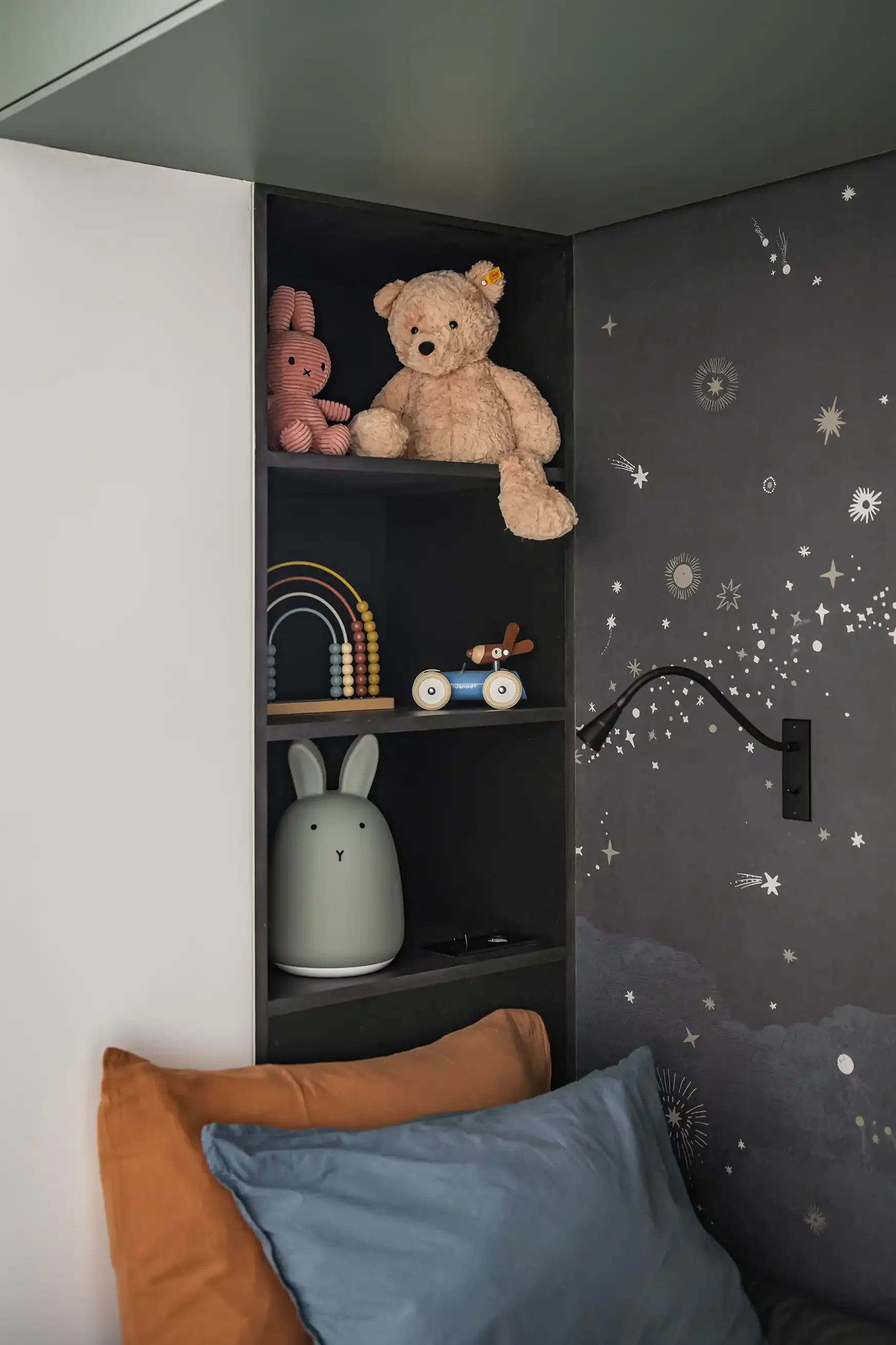 Niche de rangement noire intégrée en tête de lit avec peluches et veilleuse, à côté d'un mur décoré d'un papier peint panoramique gris aux motifs d'étoiles et de constellations.