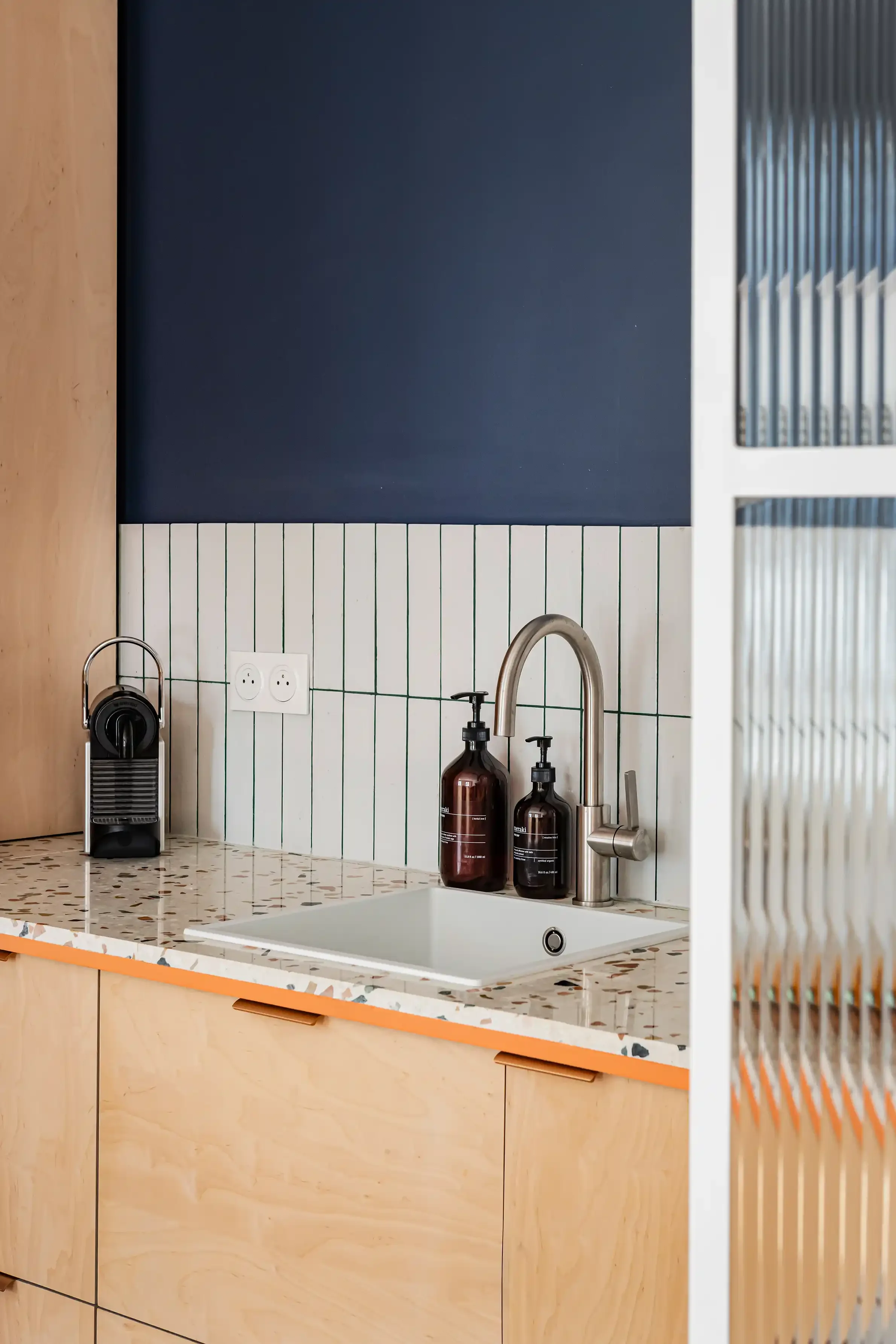 Évier de cuisine sous plan avec mitigeur noir, plan de travail en terrazzo et façades de meubles orange.