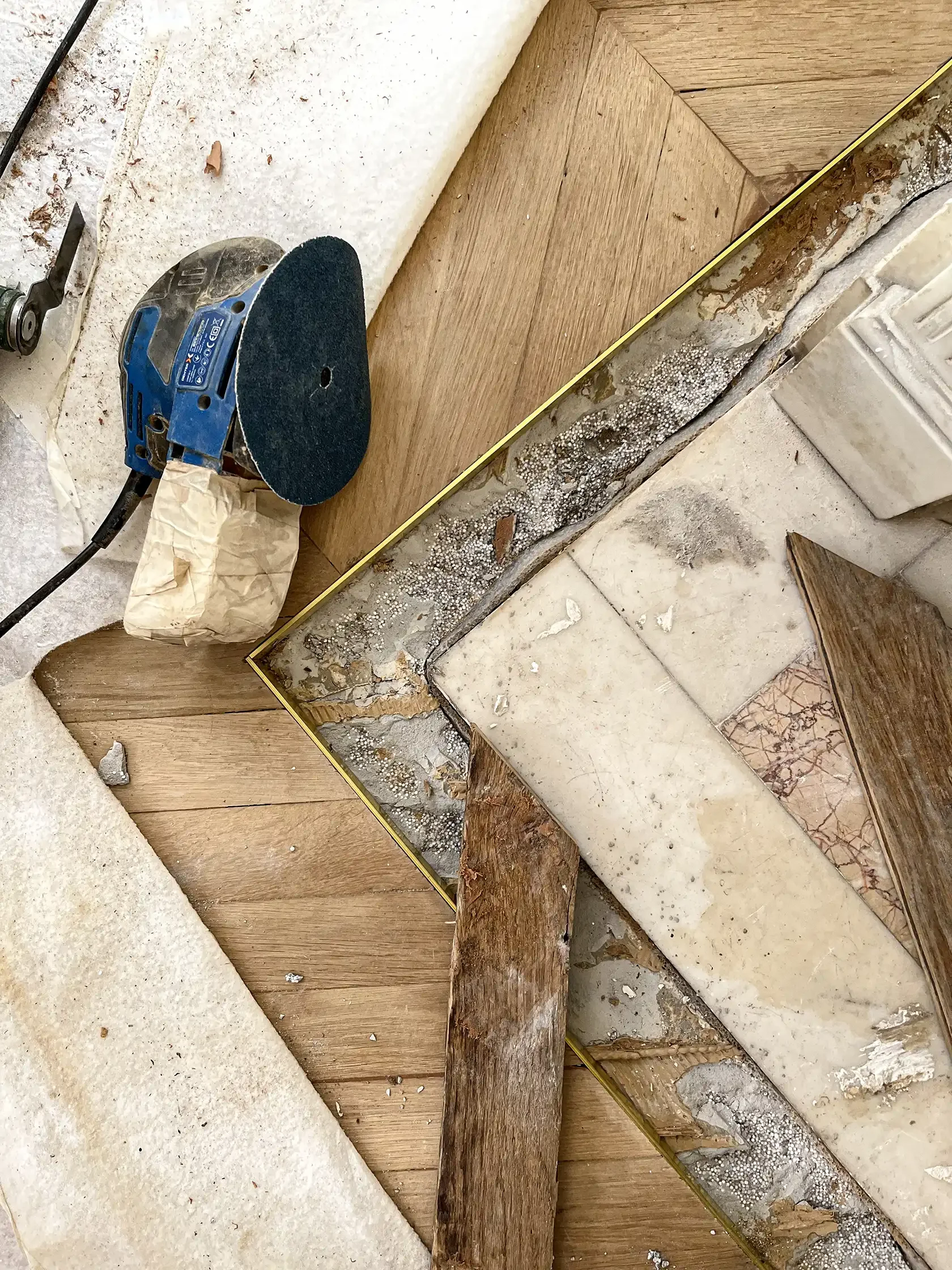 Gros plan sur la restauration d'un parquet en chêne massif point de Hongrie et d'un seuil en marbre avec une ponceuse orbitale.