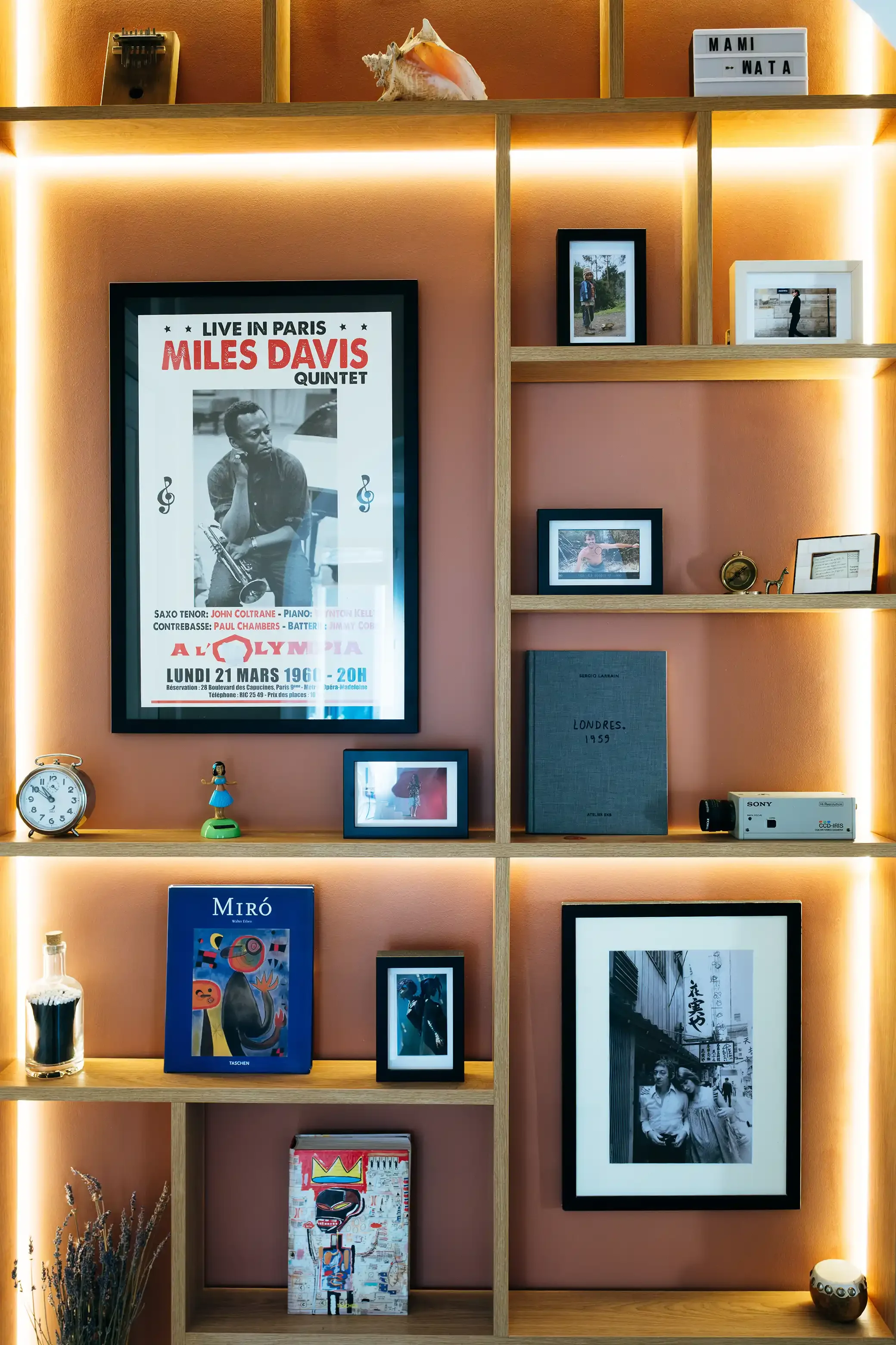 Détail d'une bibliothèque sur mesure avec rétro-éclairage LED intégré, mettant en valeur des cadres photos et des objets de collection sur un mur terracotta.