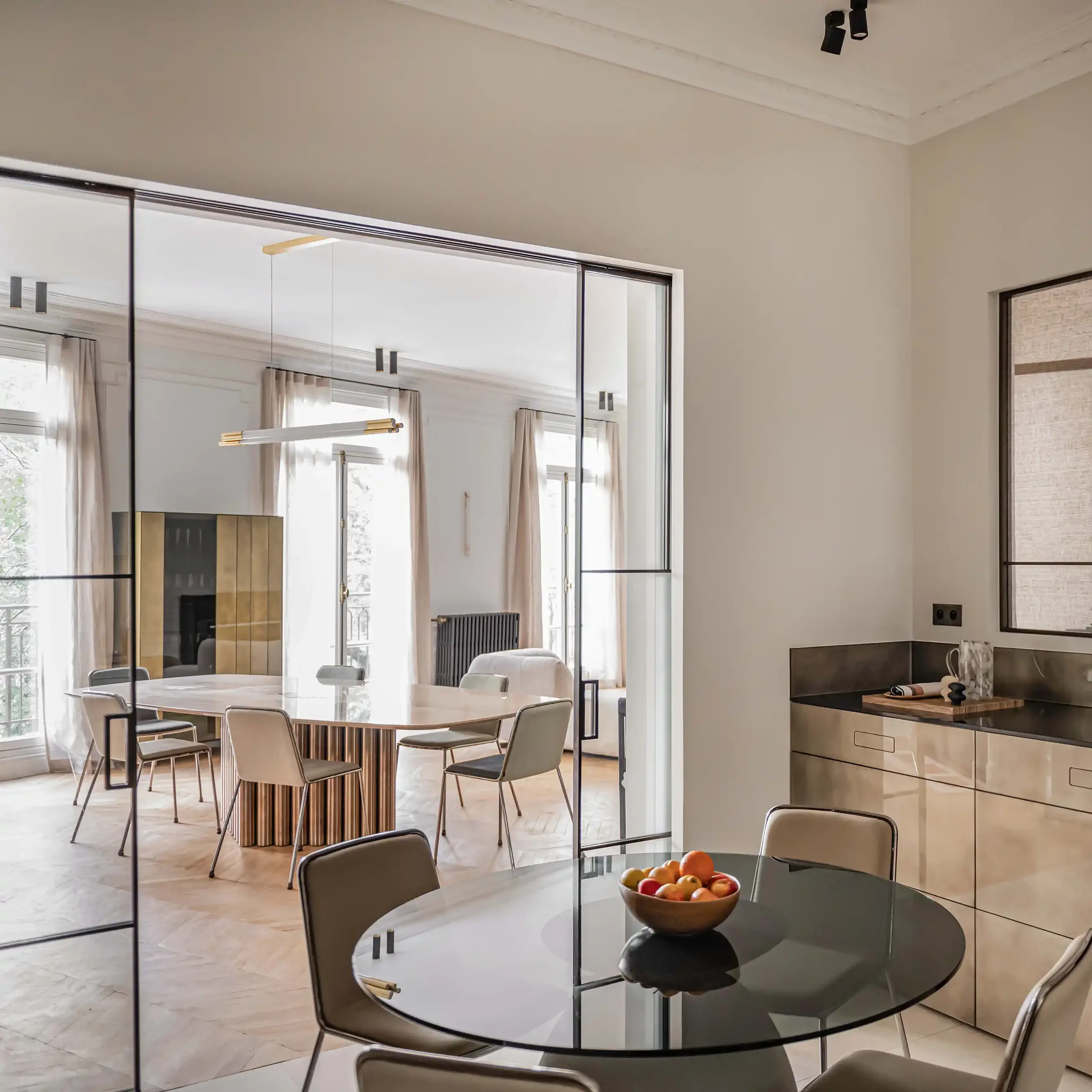 Salle à manger de luxe avec table ovale en marbre et chaises design vue à travers une verrière d'atelier noire depuis une cuisine moderne.