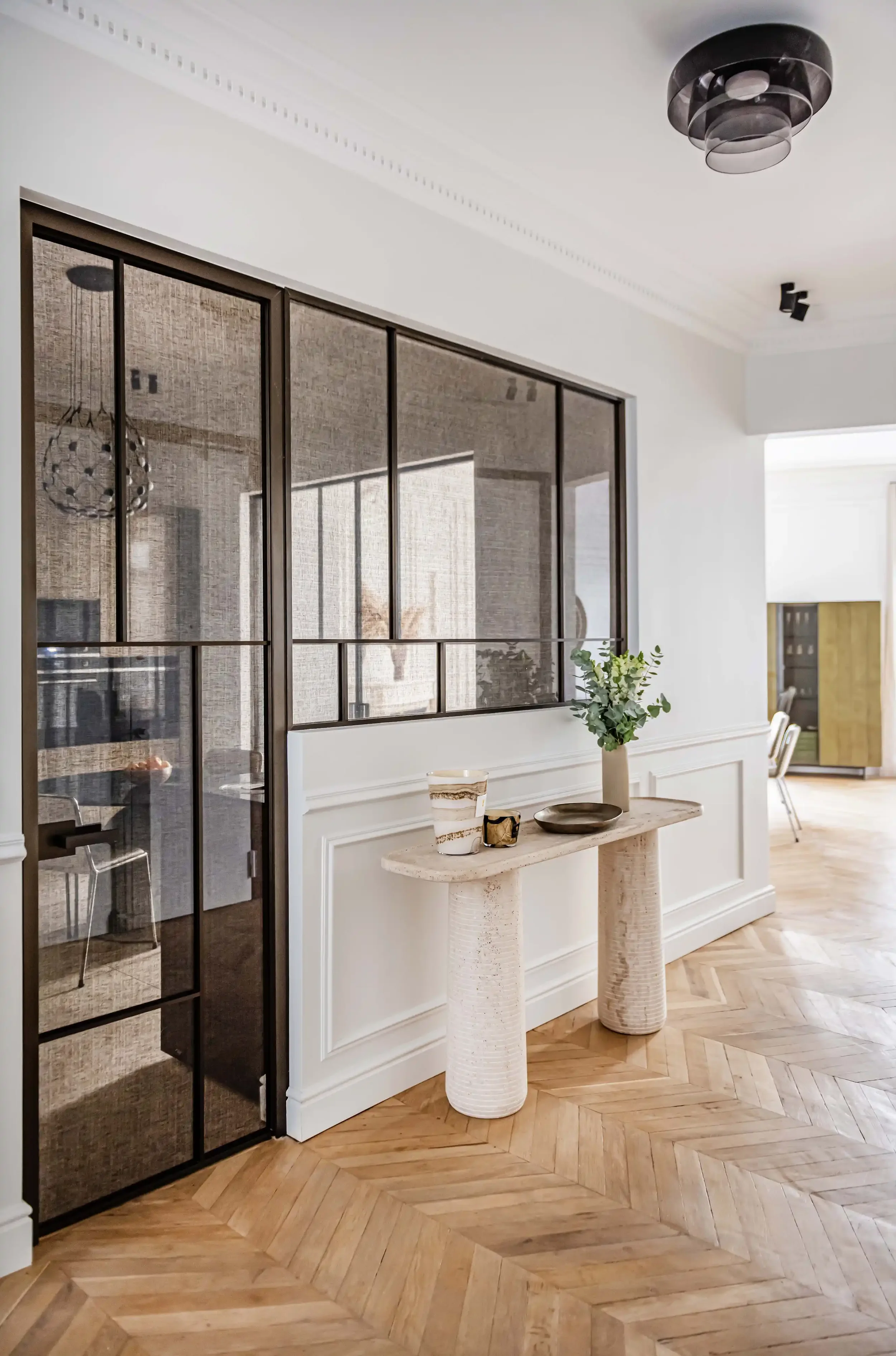 Entrée d'appartement avec verrière d'atelier en métal bronze, console fine en pierre naturelle et sol en parquet ancien.
