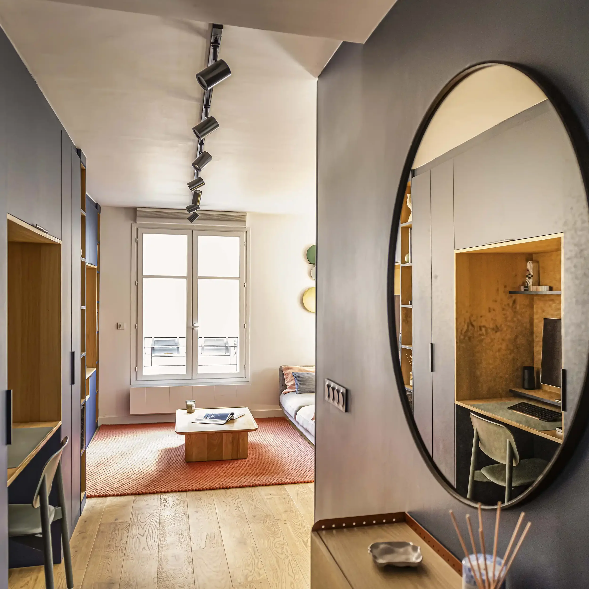 Entrée d'appartement avec grand miroir rond design, vue sur le salon et coin bureau optimisé dans le couloir.