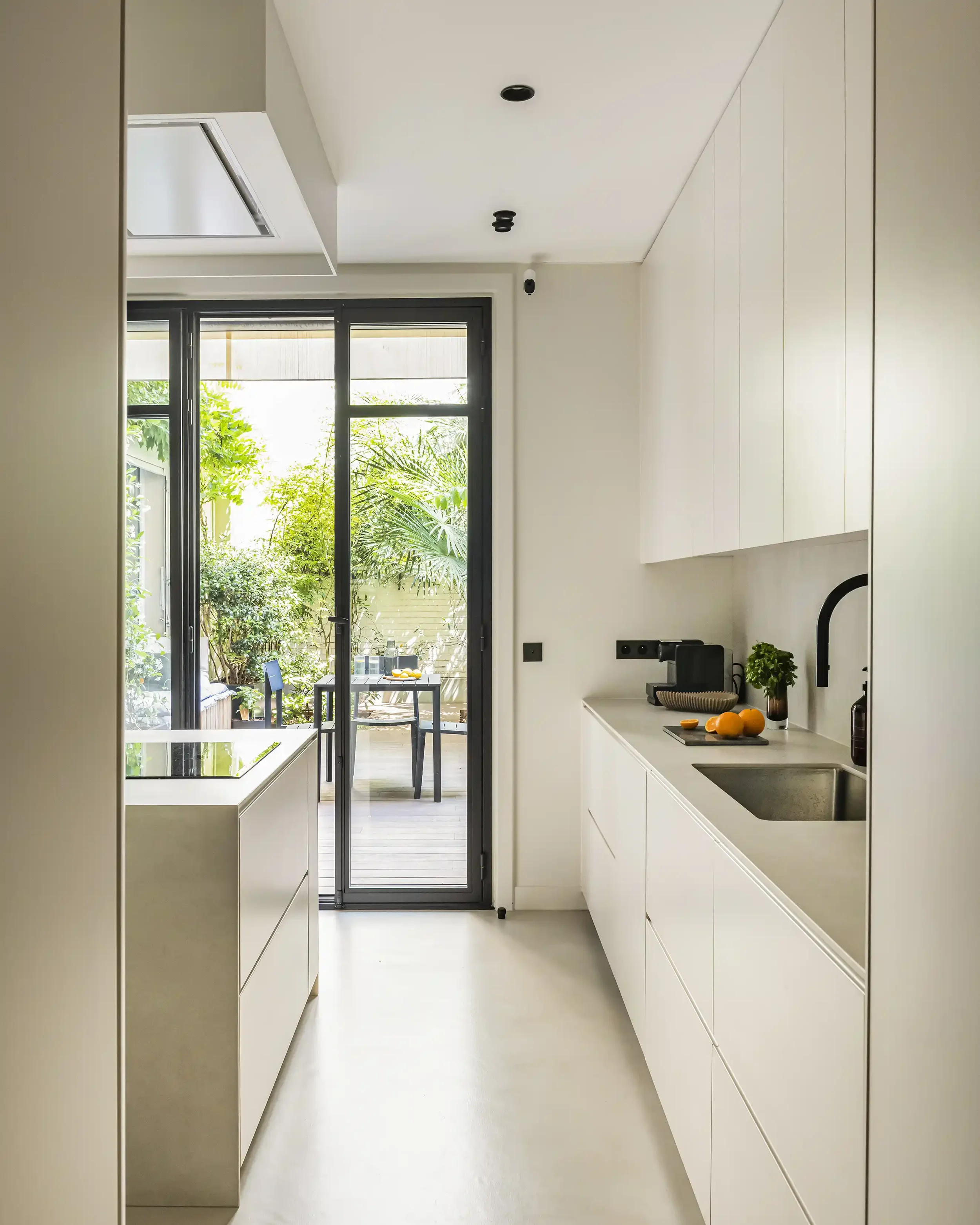 Cuisine moderne beige épurée avec îlot central offrant une vue directe sur une terrasse et un jardin arboré.