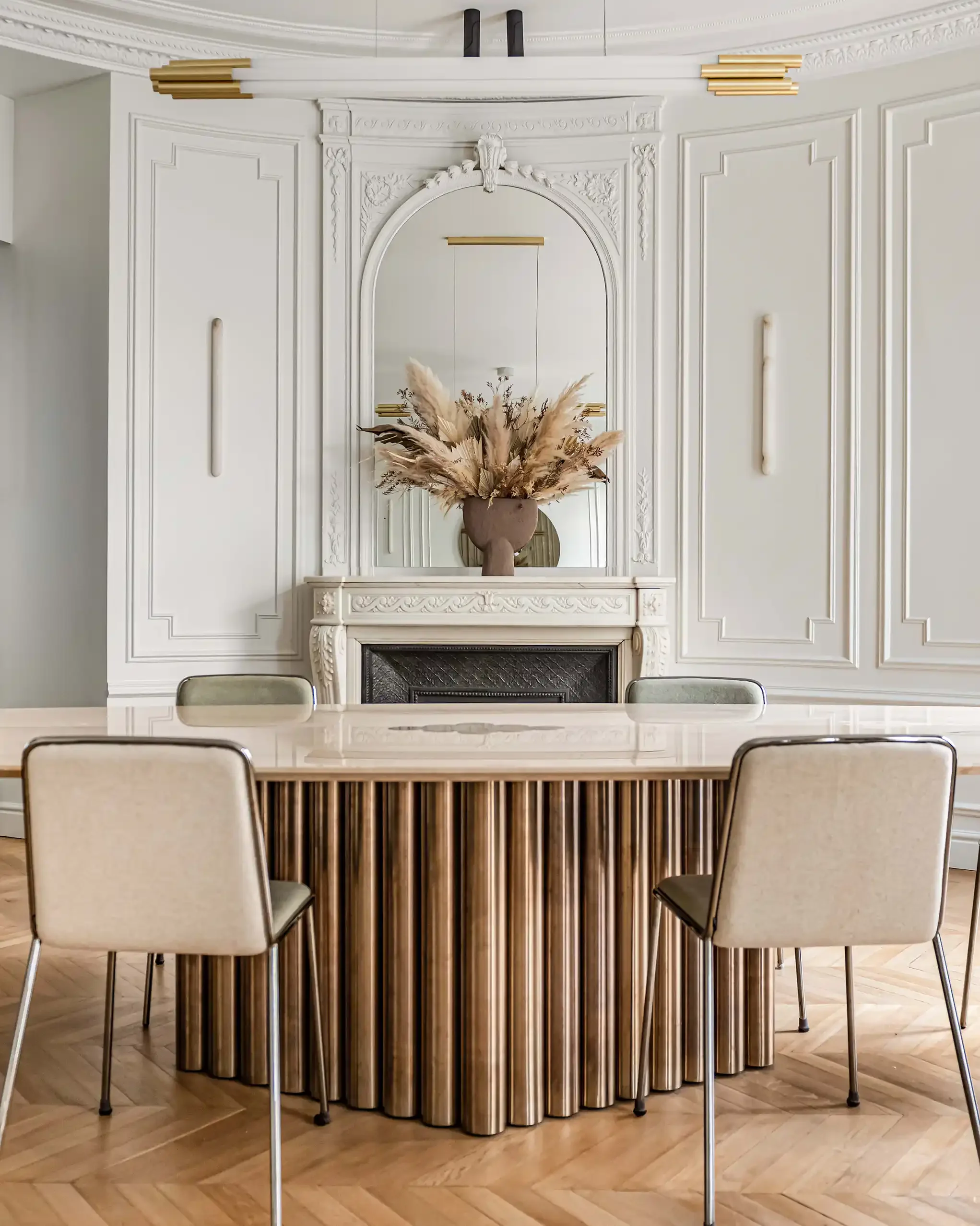Salle à manger de réception avec une grande table au piètement en tubes laiton et plateau en travertin face à une cheminée ancienne sculptée.