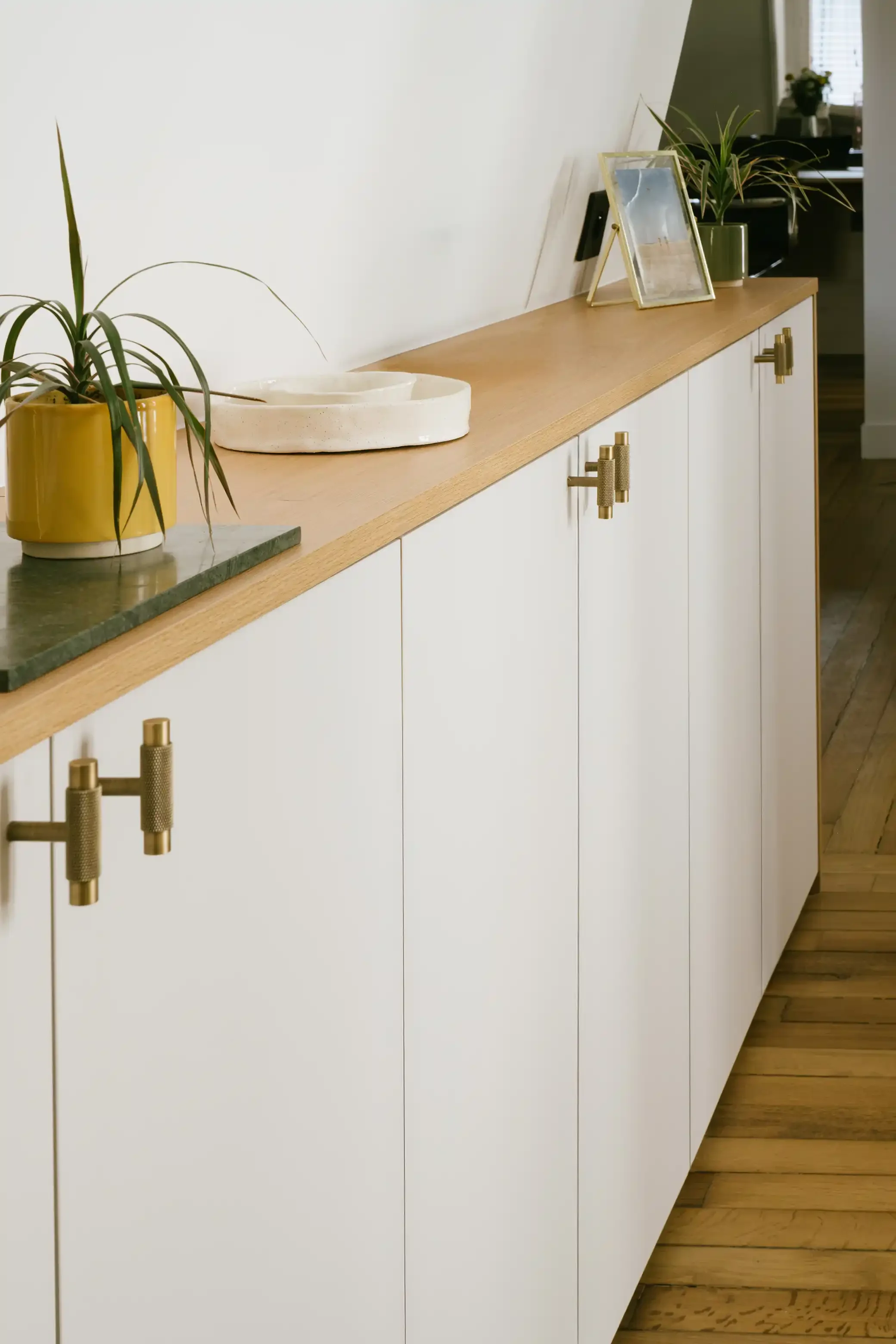 Buffet bas de salon sur-mesure au design minimaliste blanc avec plateau en chêne clair et poignées en laiton moleté, appartement parisien.