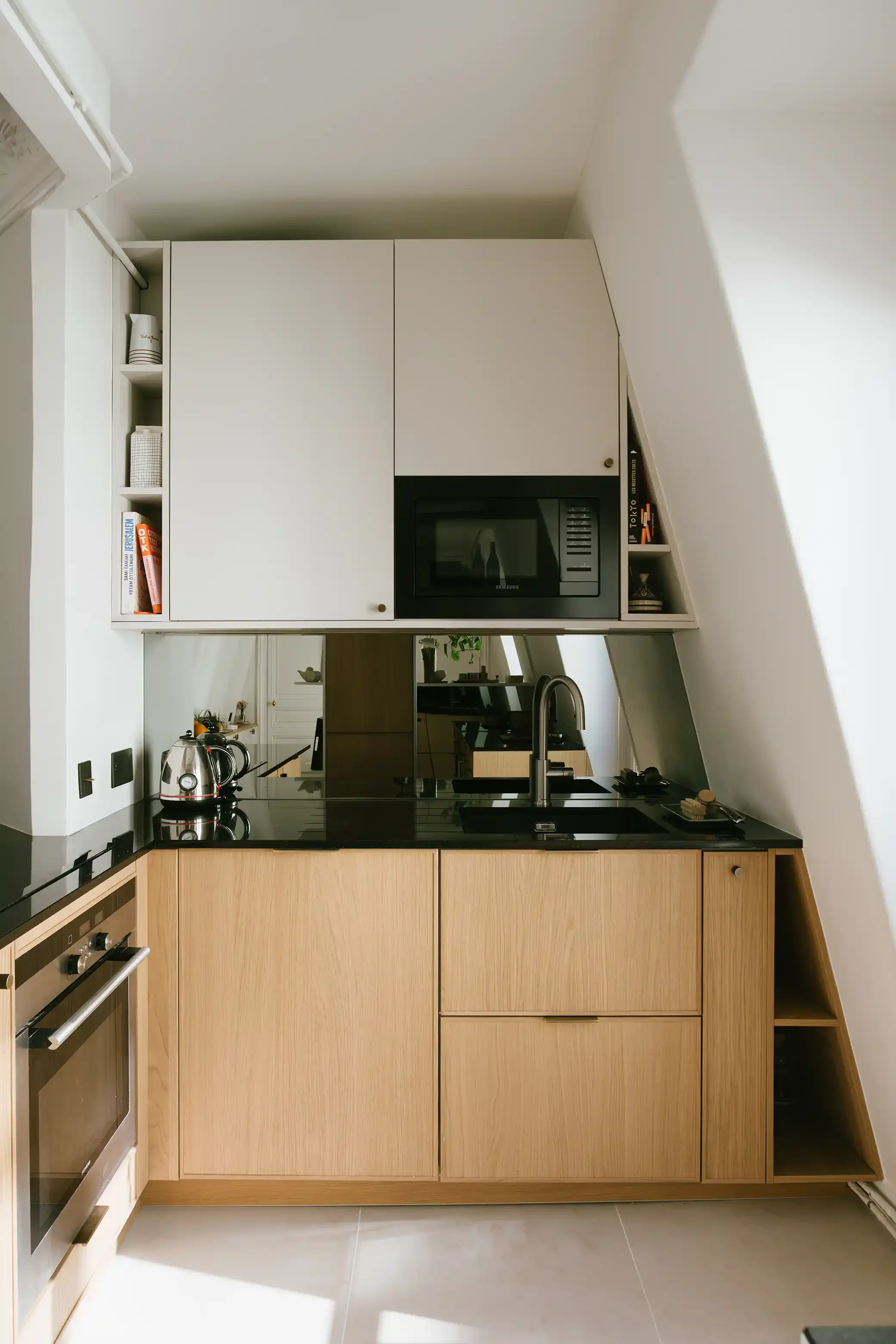 Aménagement sur mesure d'une cuisine sous pente avec façades beige et chêne, plan de travail en granit noir et étagères ouvertes optimisant l'espace.