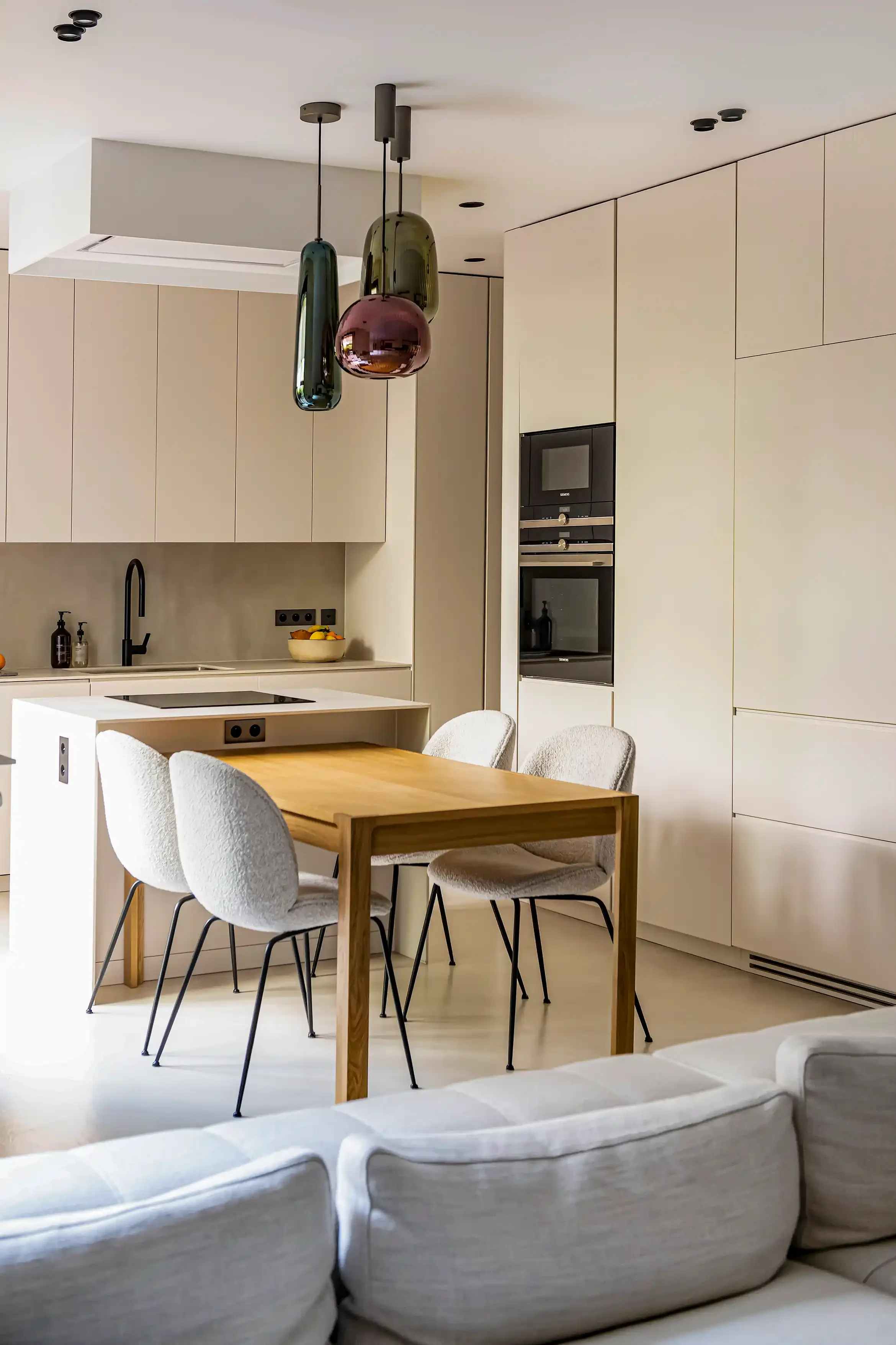 Cuisine équipée beige avec table de repas en bois clair et suspensions design en verre soufflé coloré.