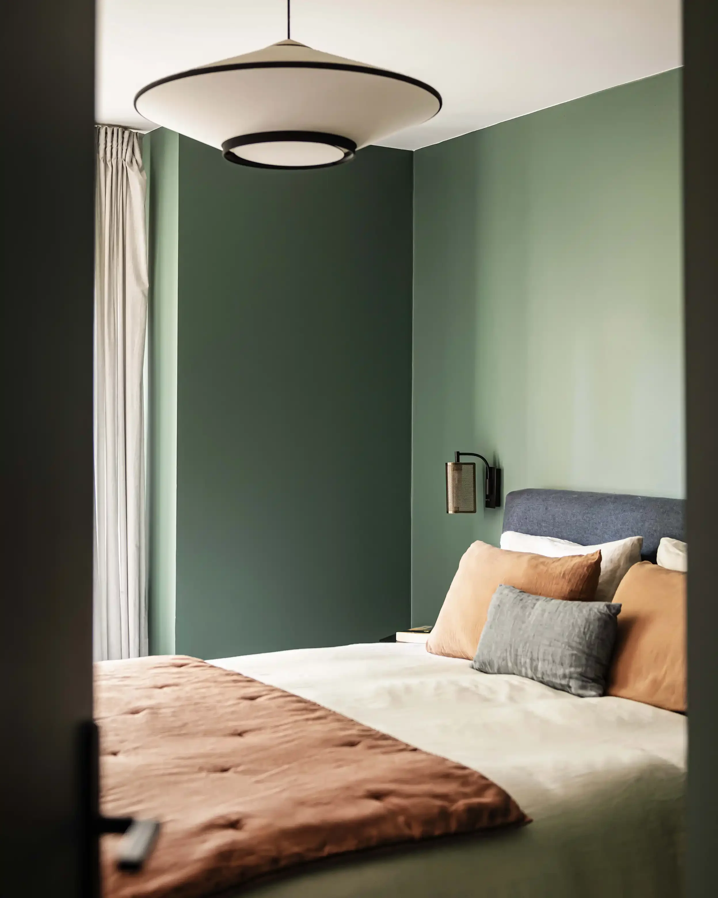 Lit douillet dans une chambre parentale aux murs vert forêt avec coussins terracotta et linge de lit blanc.