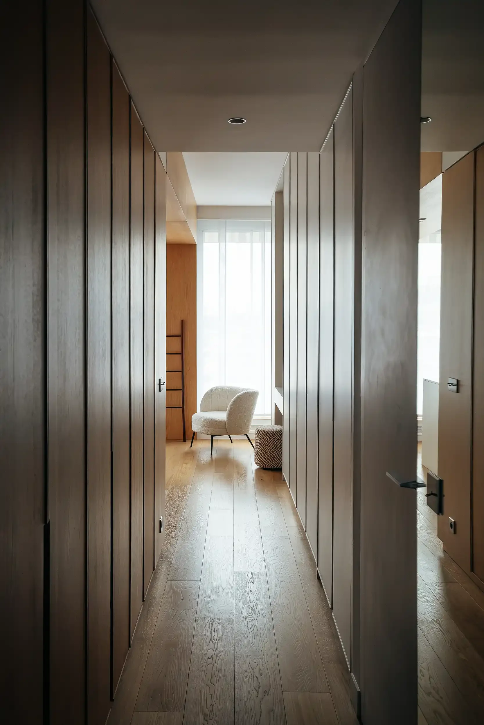 Aménagement d'un long couloir avec dressing sur mesure en bois clair et fauteuil design blanc en bouclette au fond.