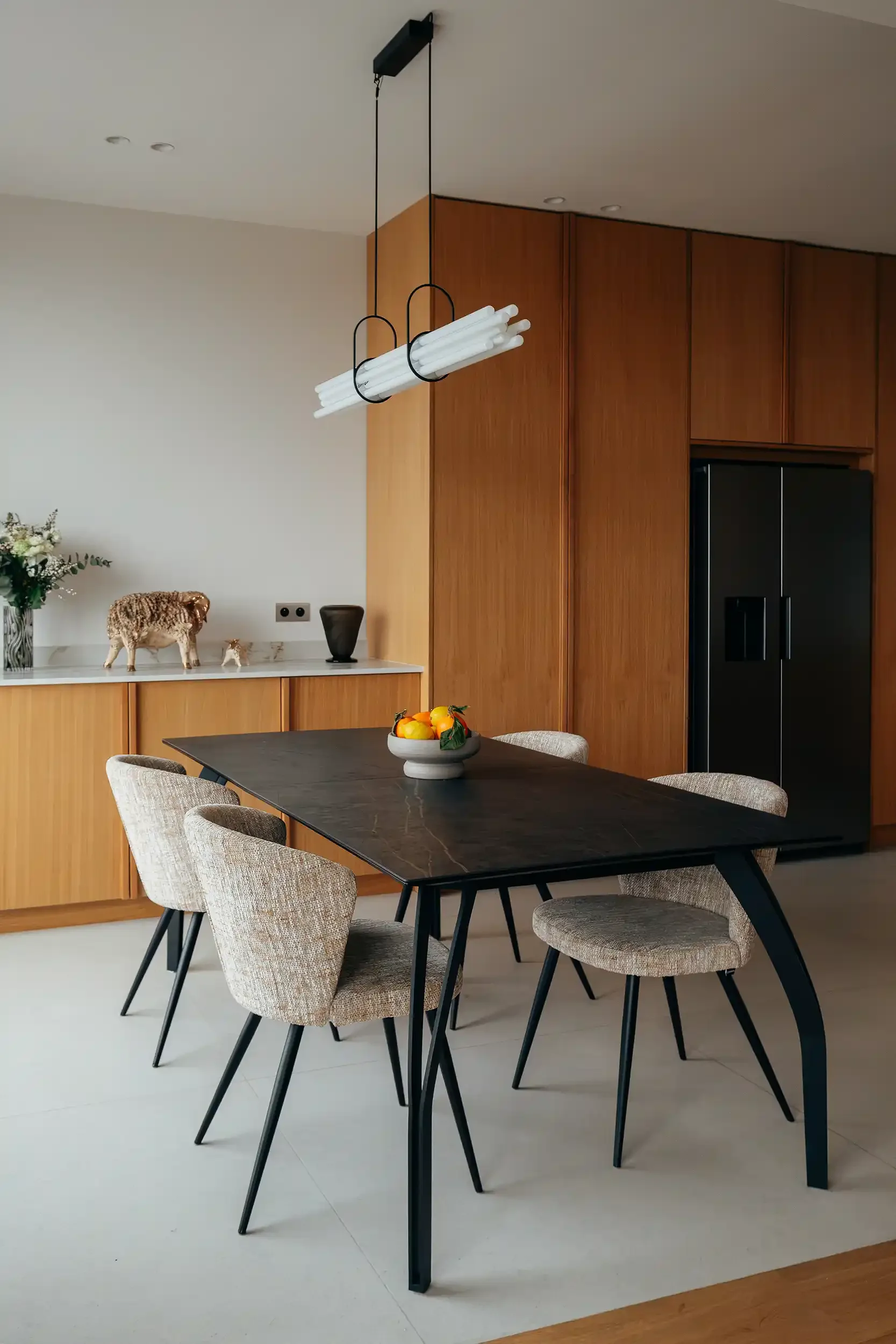 Salle à manger contemporaine avec table noire, chaises en tissu chiné et luminaire suspendu tubulaire devant une cuisine en bois.