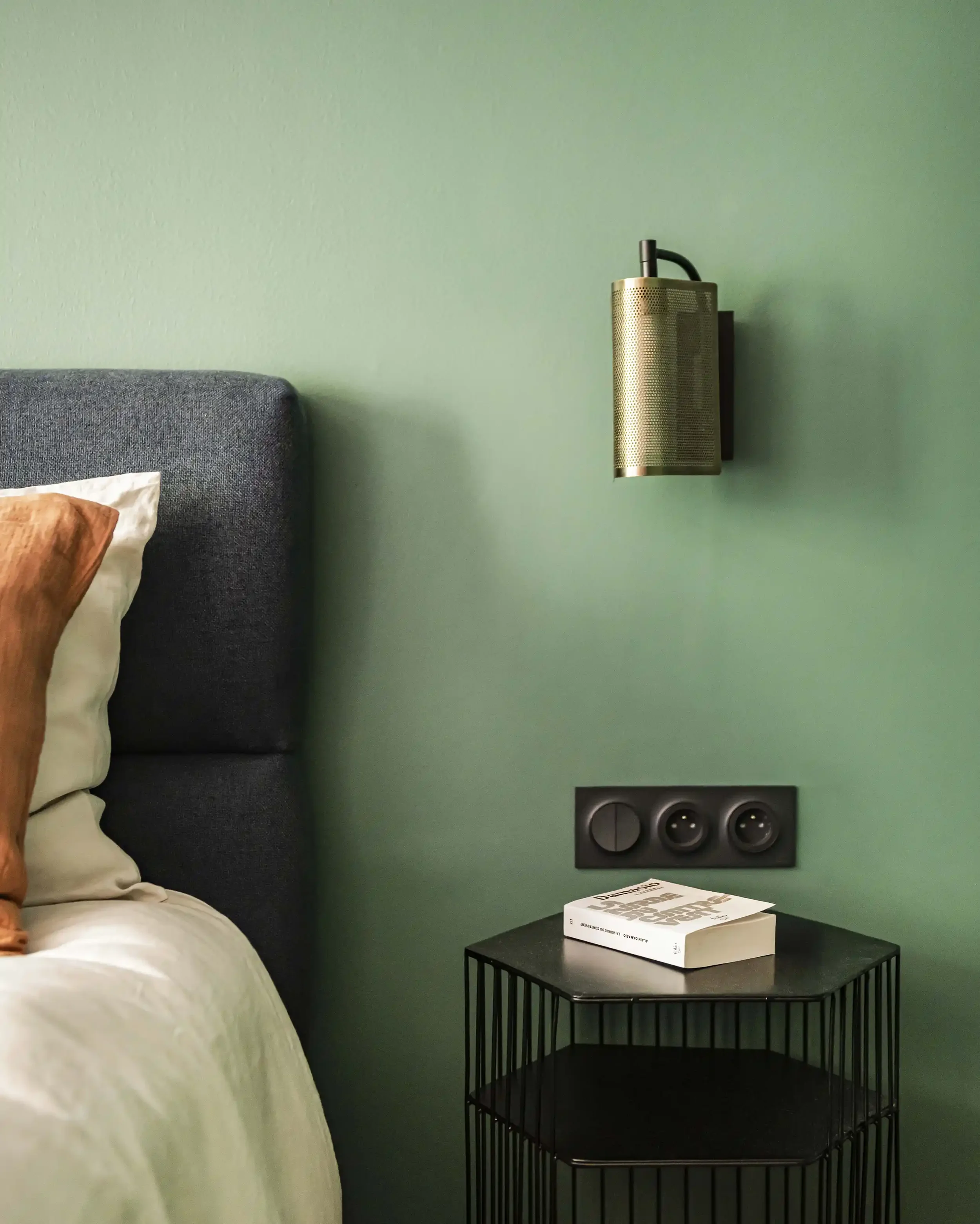 Détail luminaire de chambre avec applique en laiton perforé, interrupteur noir mat sur mur vert sauge et table de chevet design.