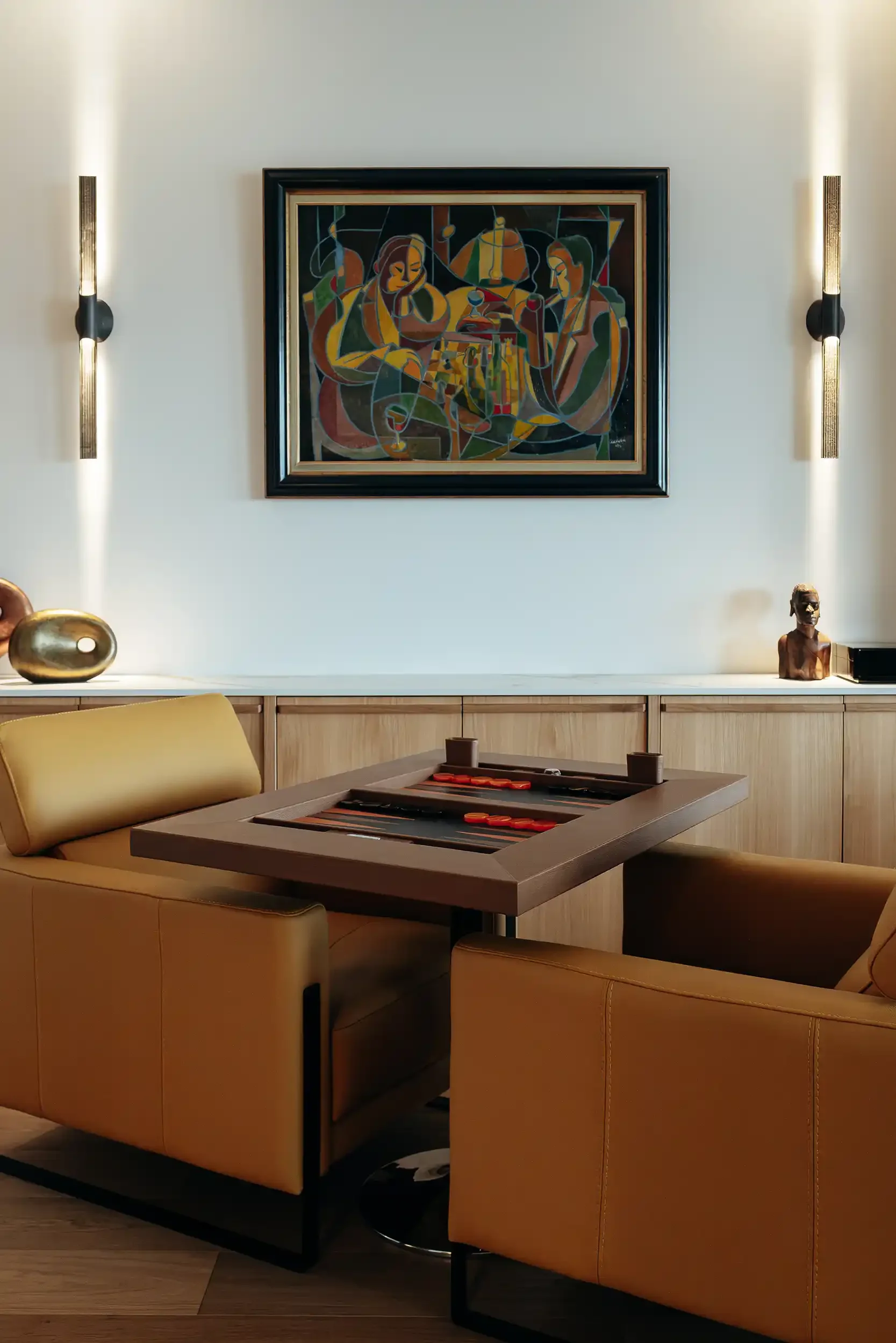 Espace de loisirs avec table de backgammon design, fauteuils en cuir camel et tableau abstrait encadré au mur.
