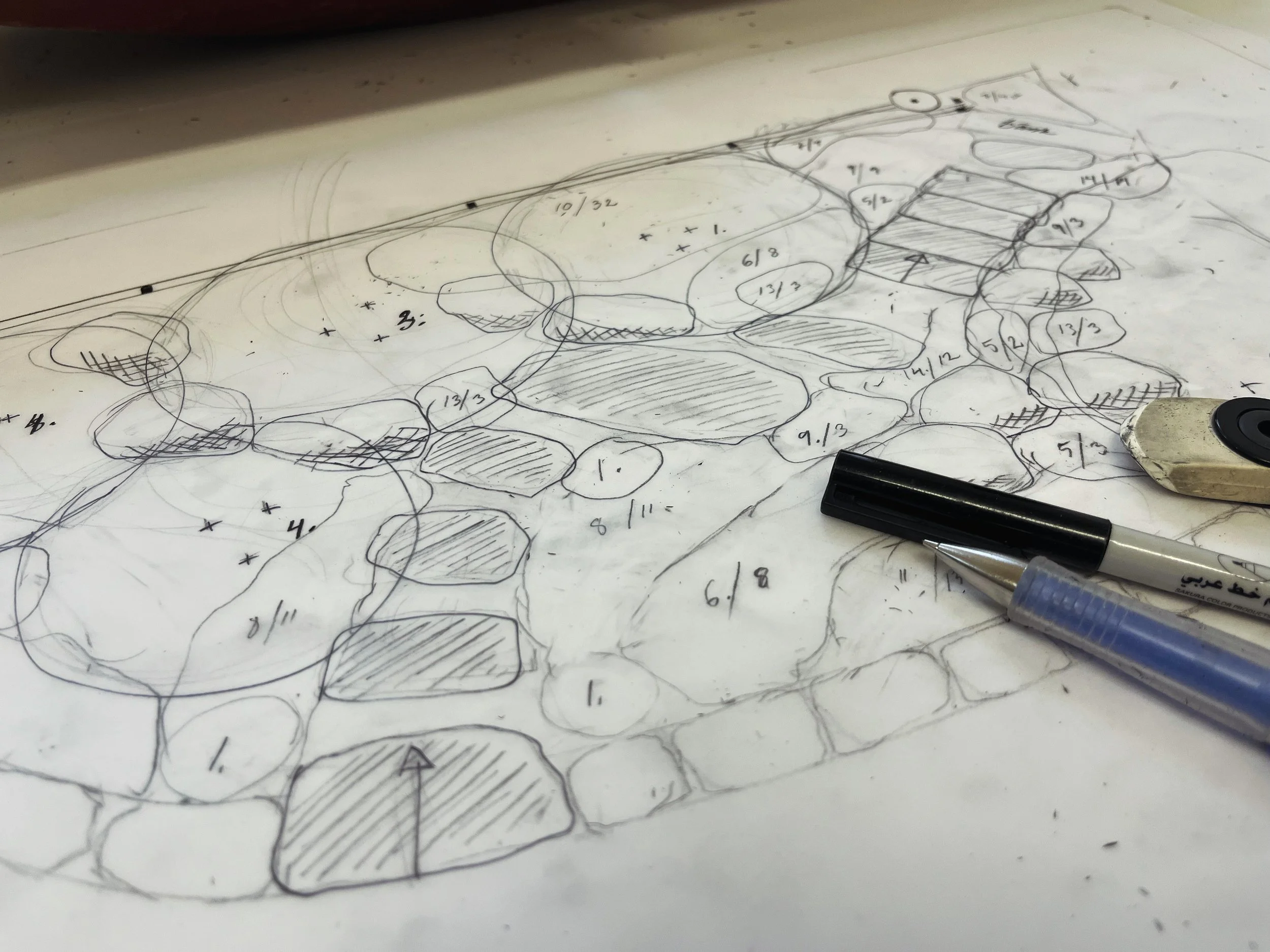 En skiss eller planritning av ett landskap eller trädgård med olika stenformationer, markerade områden, och mätningar. Två pennor och ett suddgummi ligger bredvid på papperet.