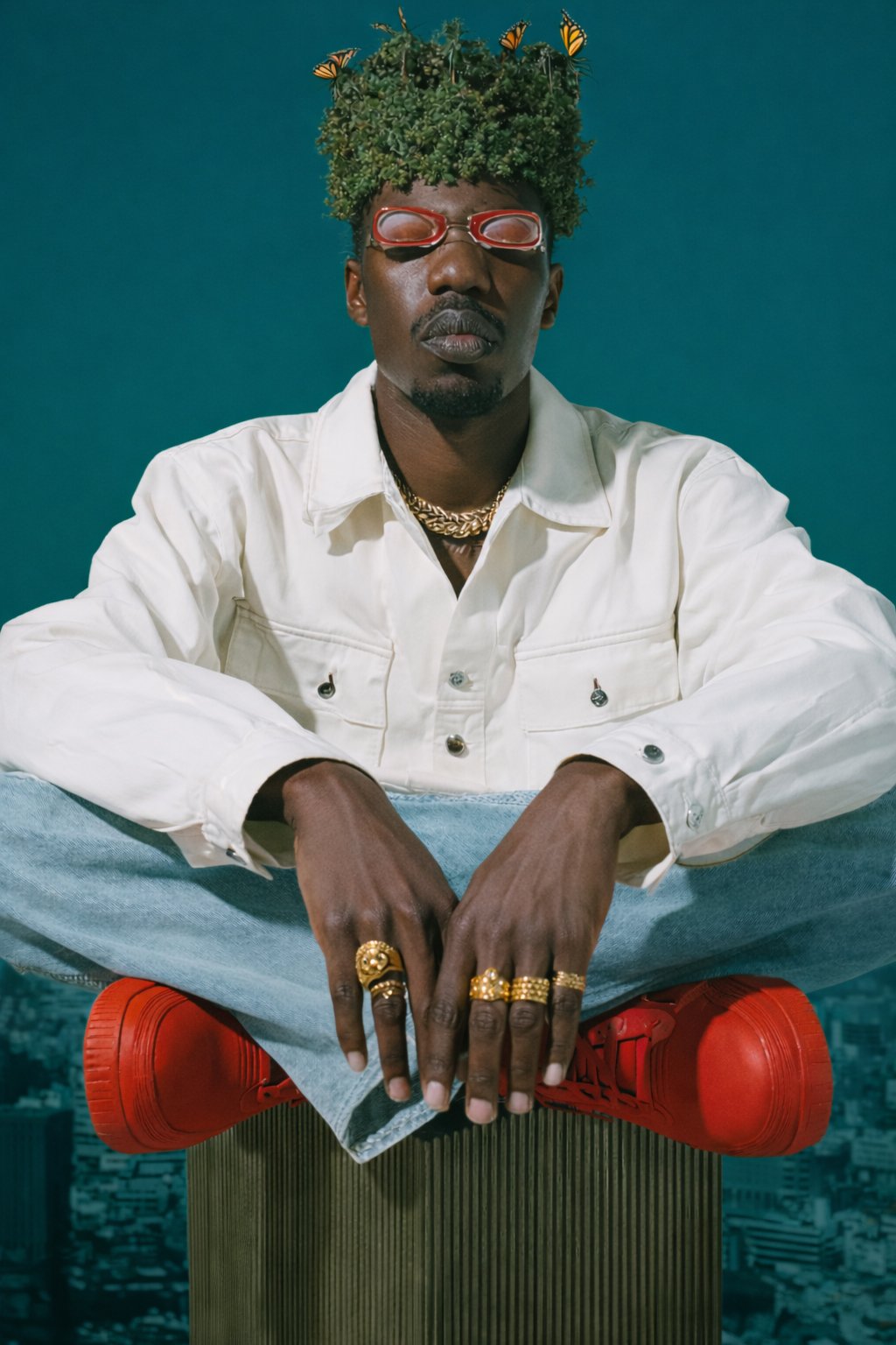Man sitting cross-legged wearing a white jacket, red shoes, gold jewelry, and sunglasses with a bushy hairstyle adorned with butterflies against a blue background.