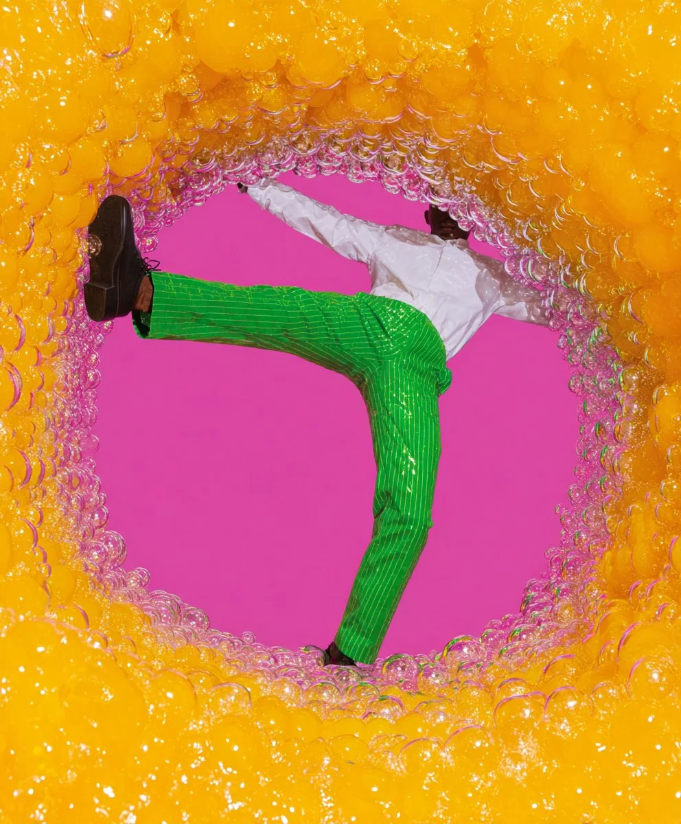 Person dressed in a white shirt and bright green striped pants, standing on one leg inside a circular tunnel of yellow and pink bubbles, against a pink background.