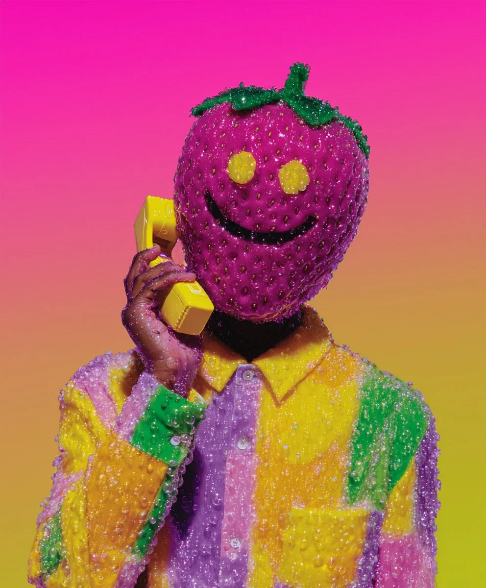Person with a strawberry for a head, smiling face, holding a yellow telephone, wearing a multicolored shirt, against a pink and orange gradient background.