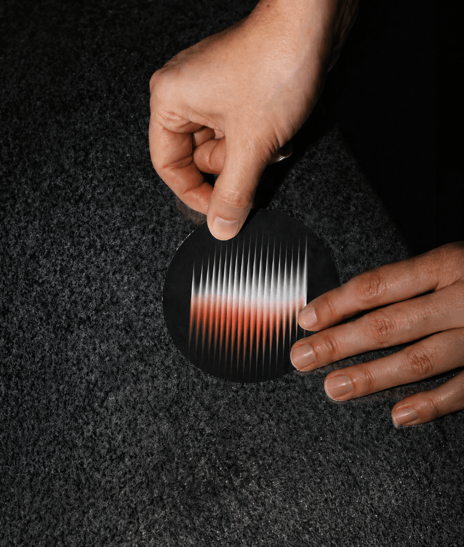 Hands applying a round sticker featuring an abstract red and white waveform graphic to a textured surface, showcasing branded merchandise for a creative agency identity.