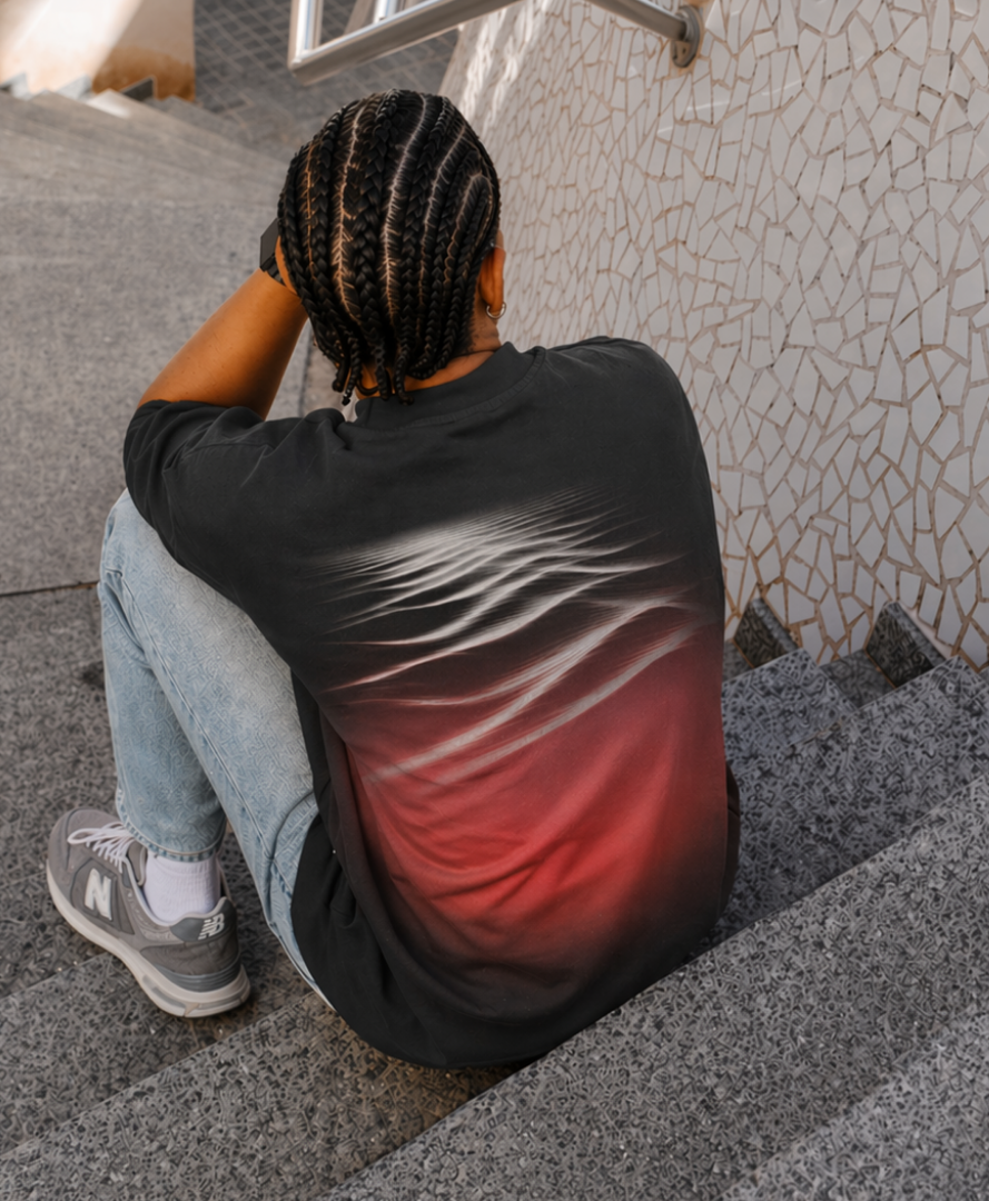 Person sitting on concrete steps wearing a black T-shirt featuring an abstract red and white motion graphic across the back, showcasing branded apparel for a creative agency identity.