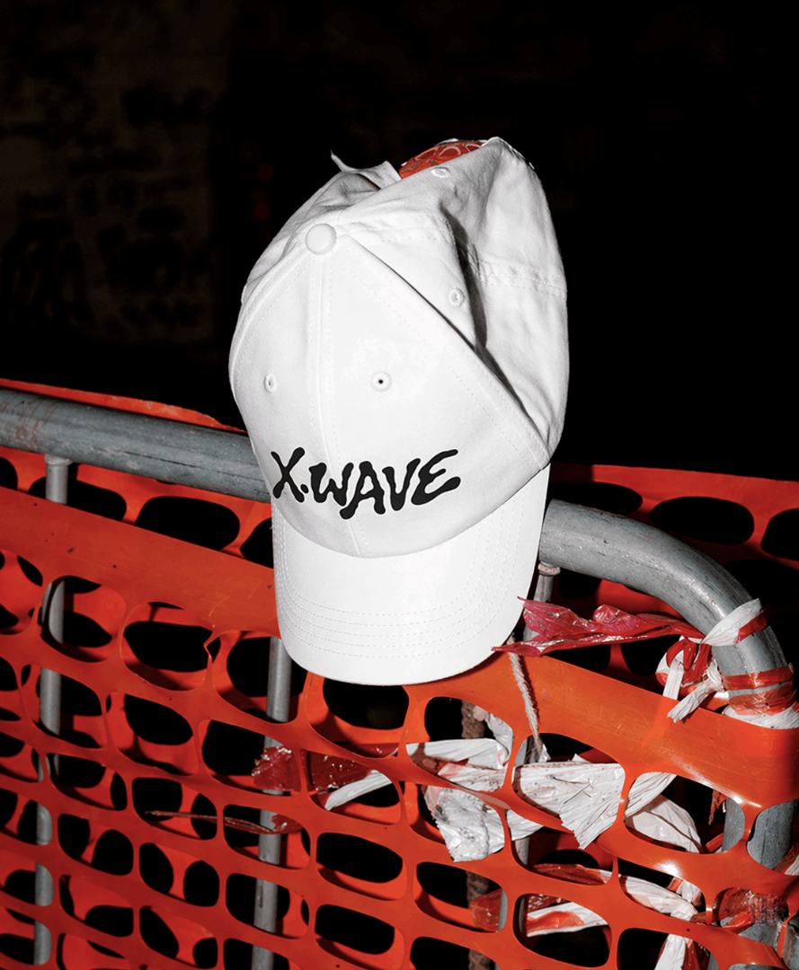 White branded baseball cap featuring a custom logo, photographed on orange construction fencing as part of a creative agency's merchandise and brand identity system.