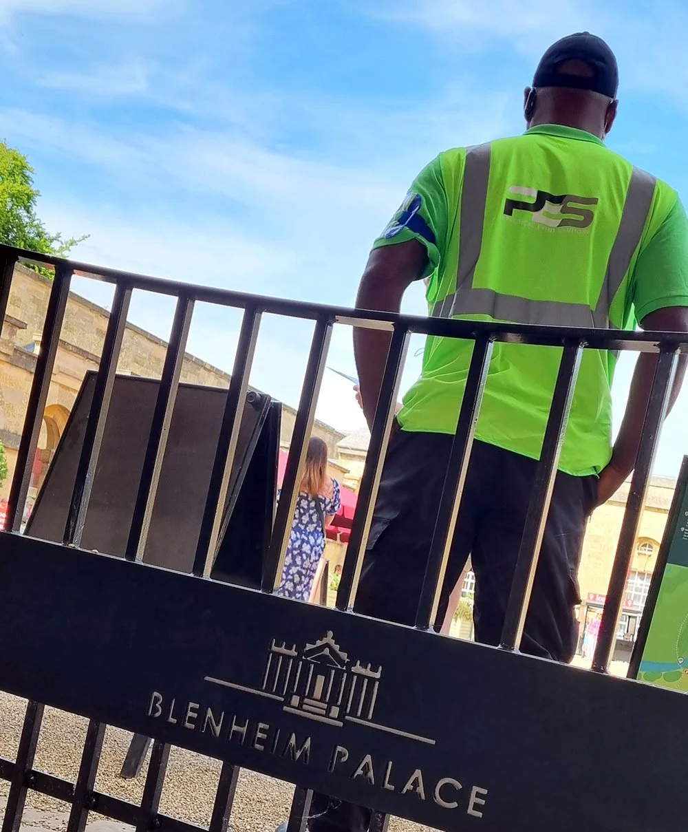 Man wearing a bright green safety vest with 'PES' logo, standing on a street near Blenheim Palace, with a woman in a floral dress in the background and a clear blue sky overhead.