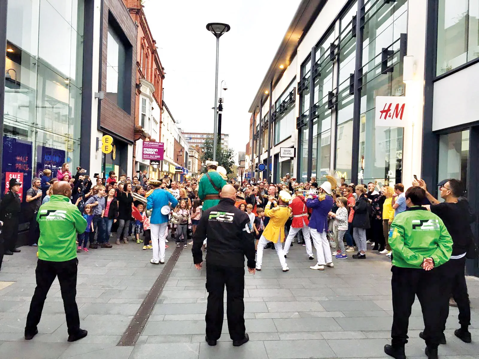 A lively street scene with a crowd of people gathered, some dressed in colorful costumes and uniforms, some taking photos or videos. Several officers are present, and tall buildings with glass storefronts and signage, including H&M, line the street.