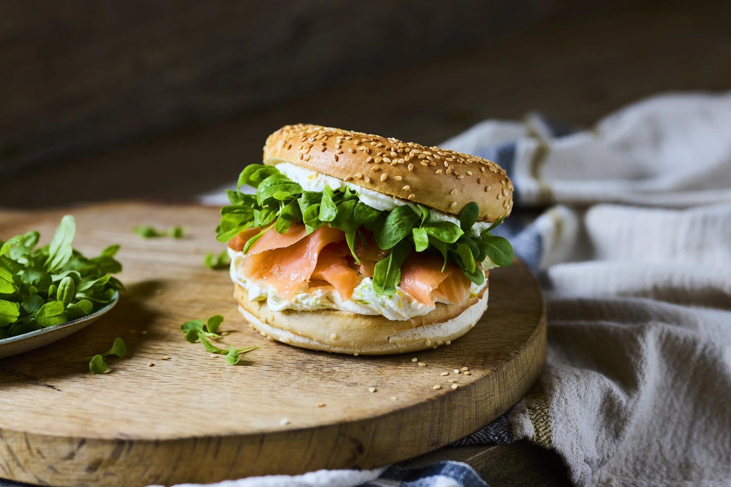 Sandwich with smoked salmon, fresh greens, and creamy spread on a sesame seed bun, placed on a wooden board.