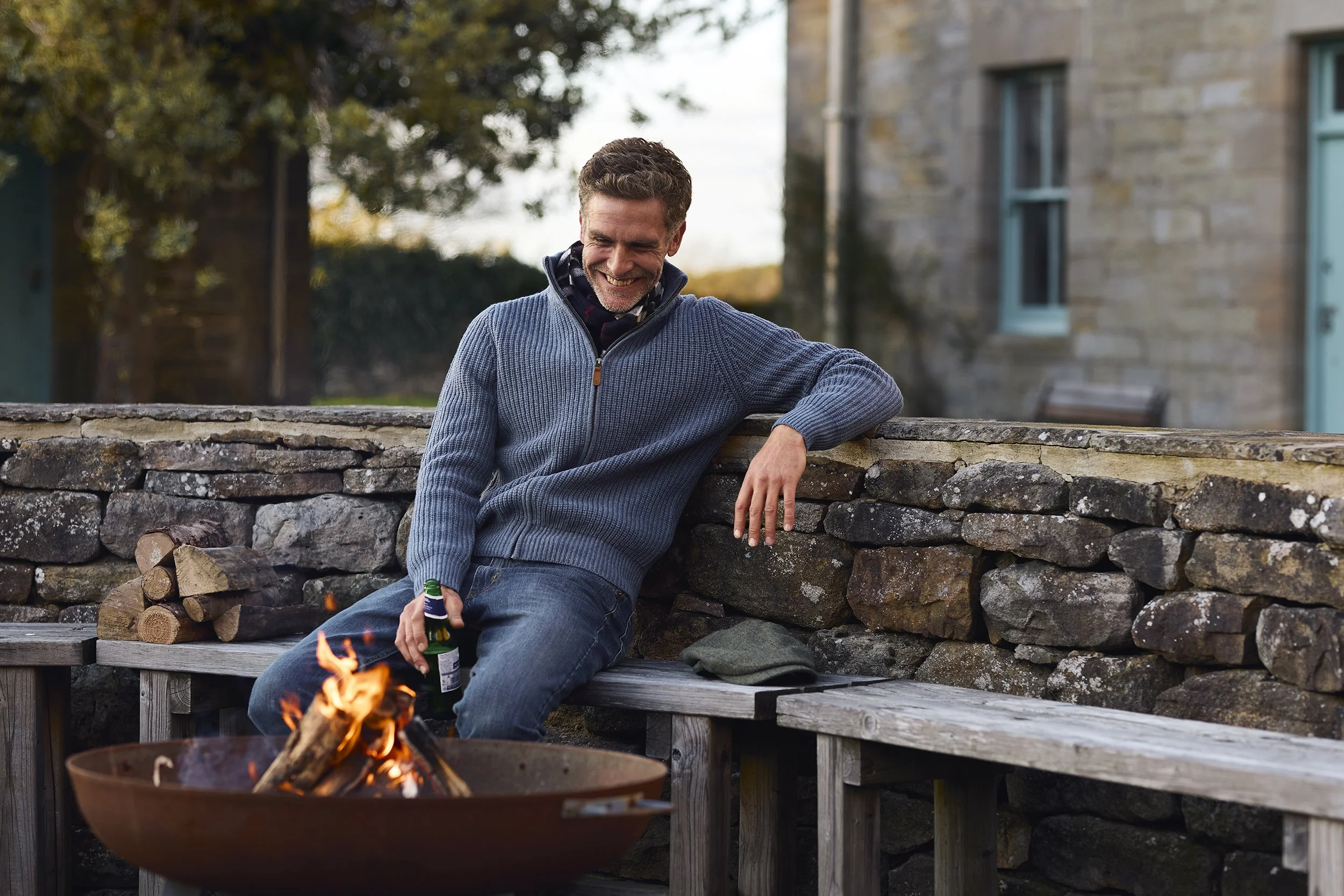 A man sitting on a wooden bench by a stone wall, holding a beer, smiling, next to a small outdoor fire.