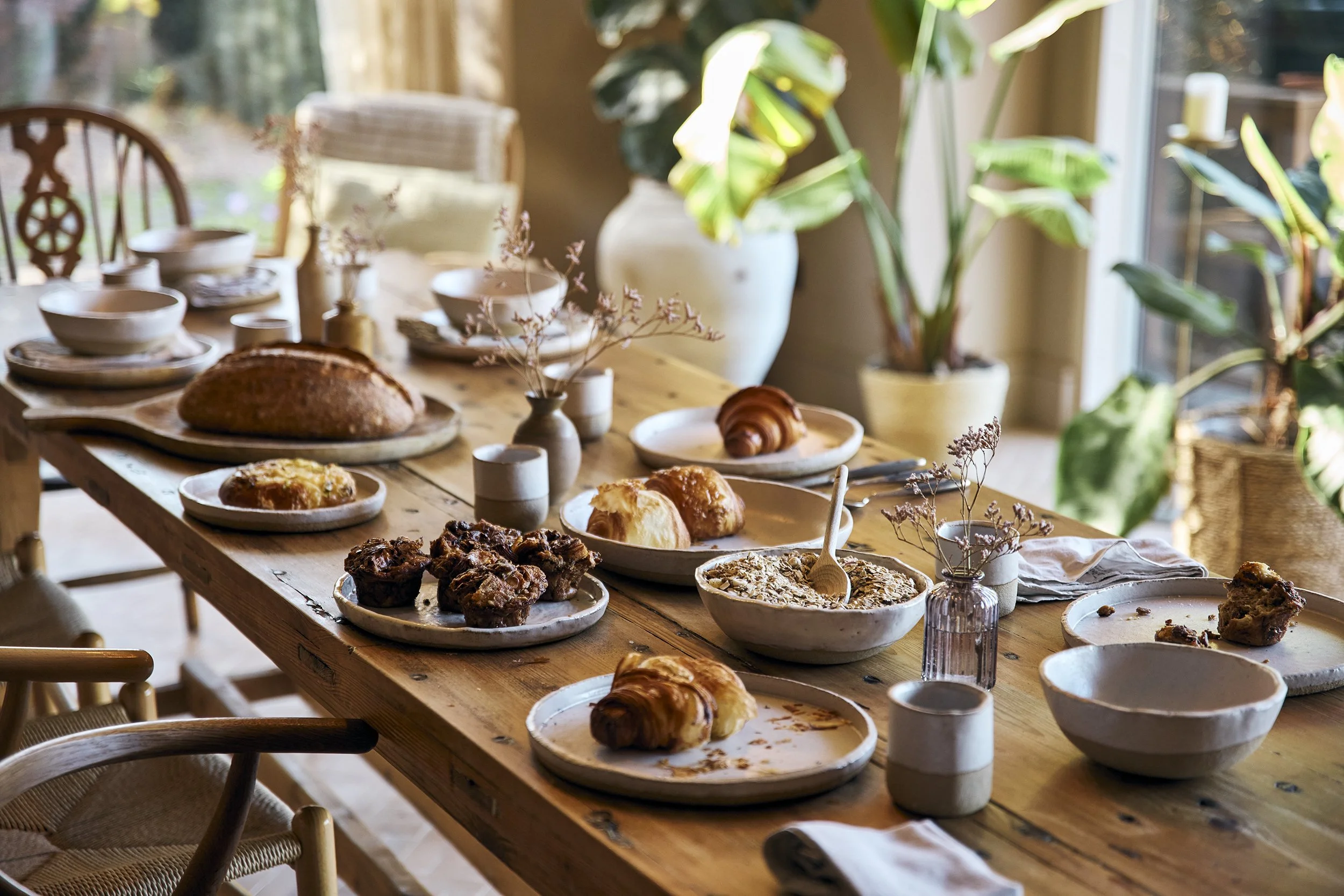 A wooden dining table set for breakfast with various baked goods and bowls, surrounded by chairs and large indoor plants in the background.