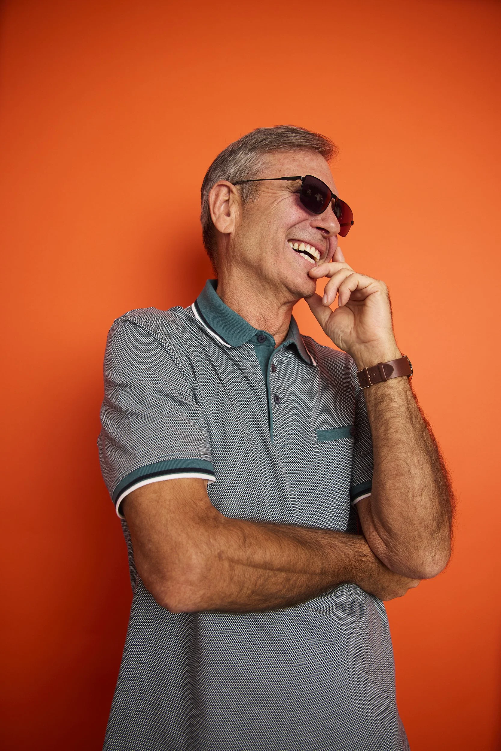 A smiling middle-aged man wearing sunglasses and a gray polo shirt with teal accents, standing against an orange background, with his arms crossed and hand touching his face.