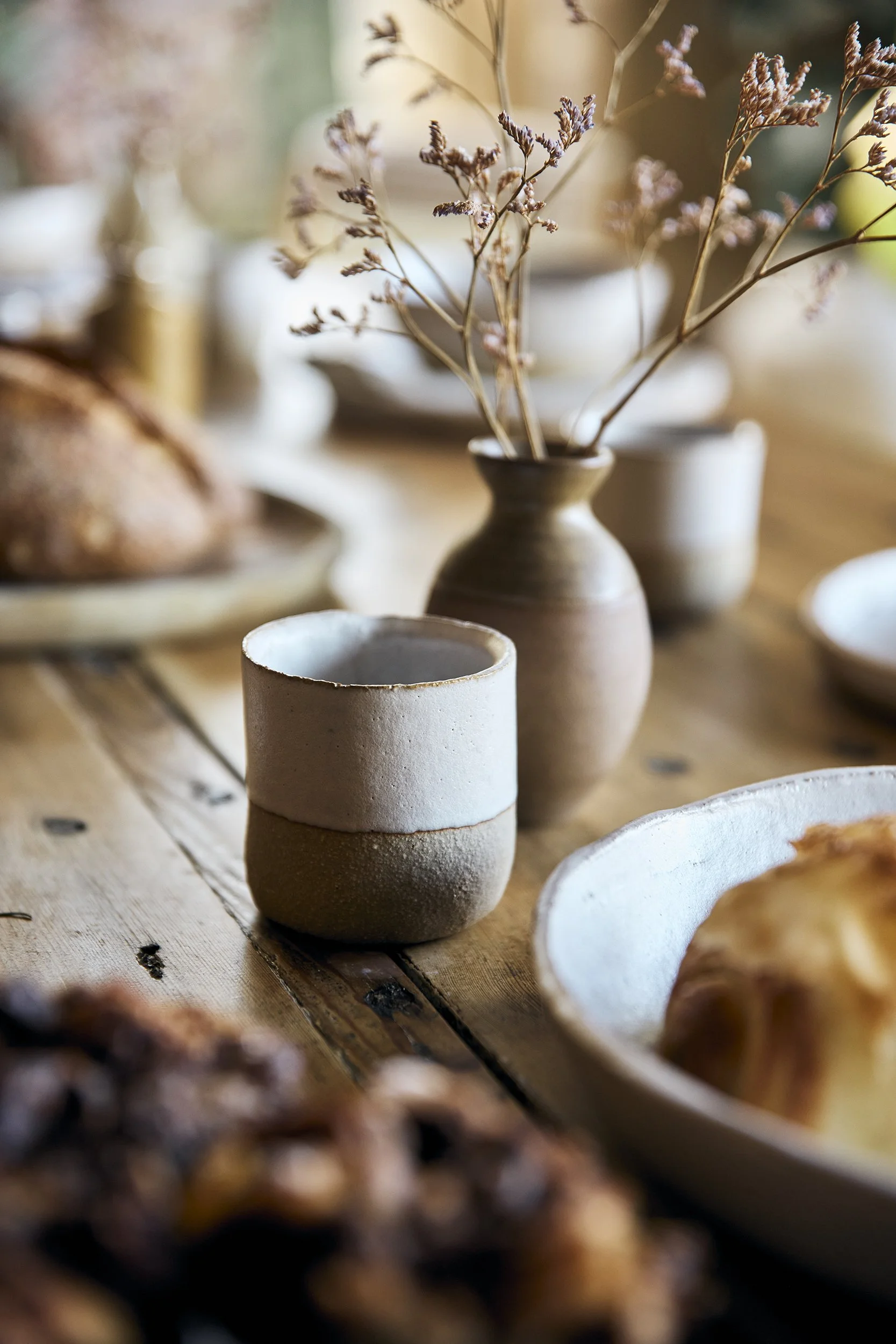 A close-up of a rustic table set with ceramic cups, a small vase with dried flowers, a bowl with baked bread, and other dishes in a warm, cozy setting.