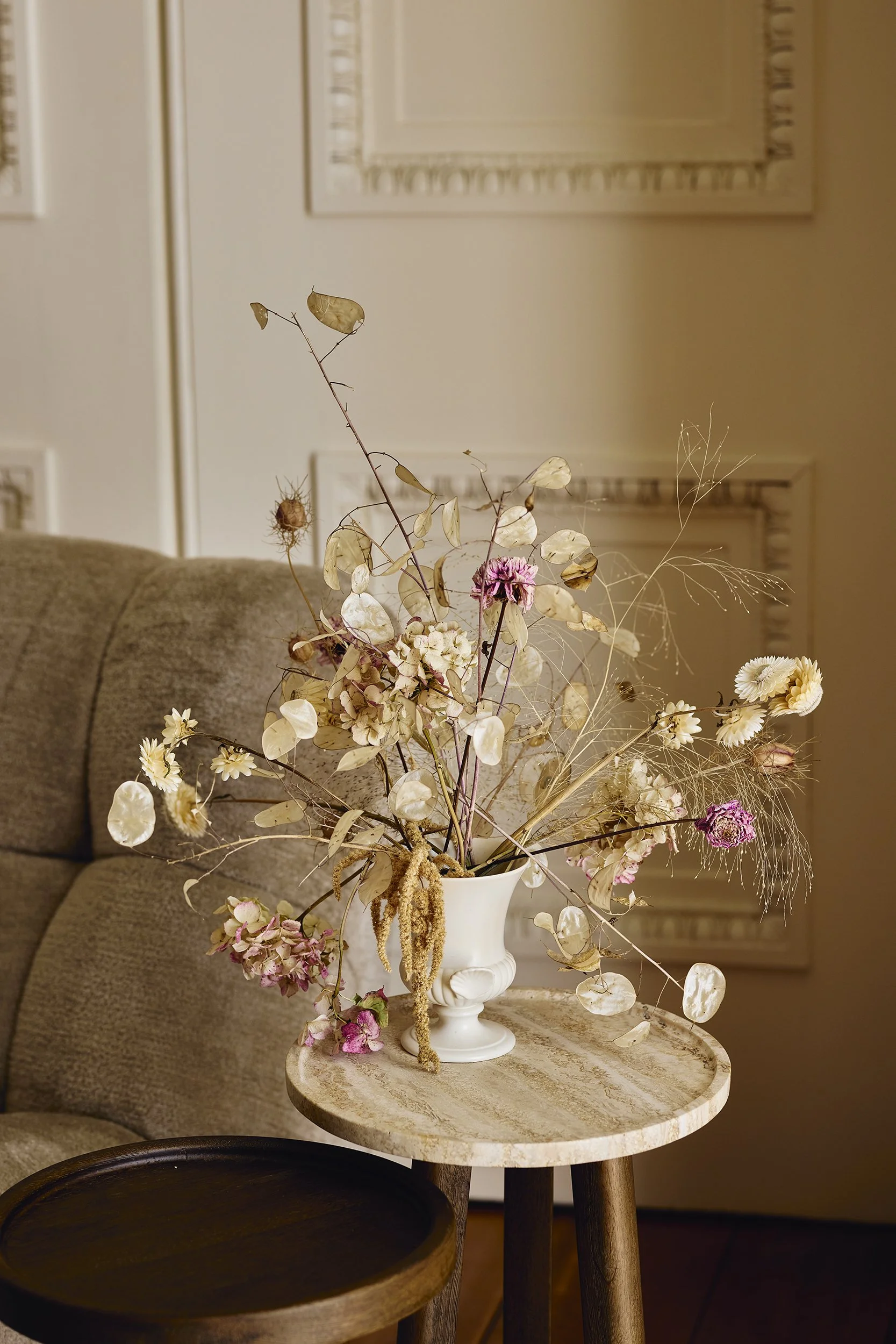 A beige couch, a small round wooden table, and a white vase with dried flowers including pink, white, and beige blooms, on a light-colored table.