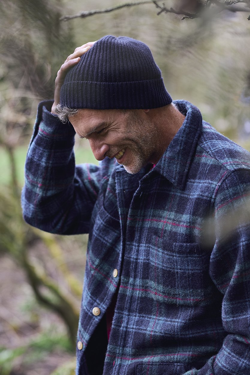 A man wearing a blue beanie and a plaid flannel shirt, smiling and holding his head with one hand, outdoors in a natural setting.