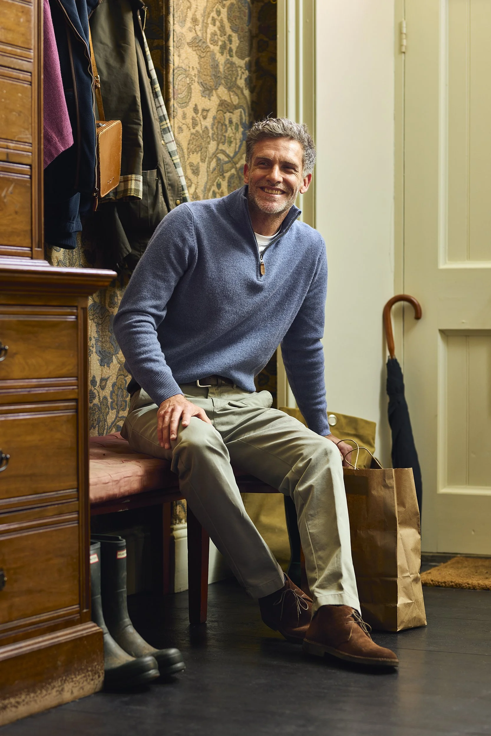 A man with gray hair and a beard sitting on a pink cushioned bench in a hallway, smiling. He's dressed in a blue quarter-zip sweater, beige pants, and brown shoes, holding a brown paper shopping bag. Behind him are hanging jackets, a floral wallpaper