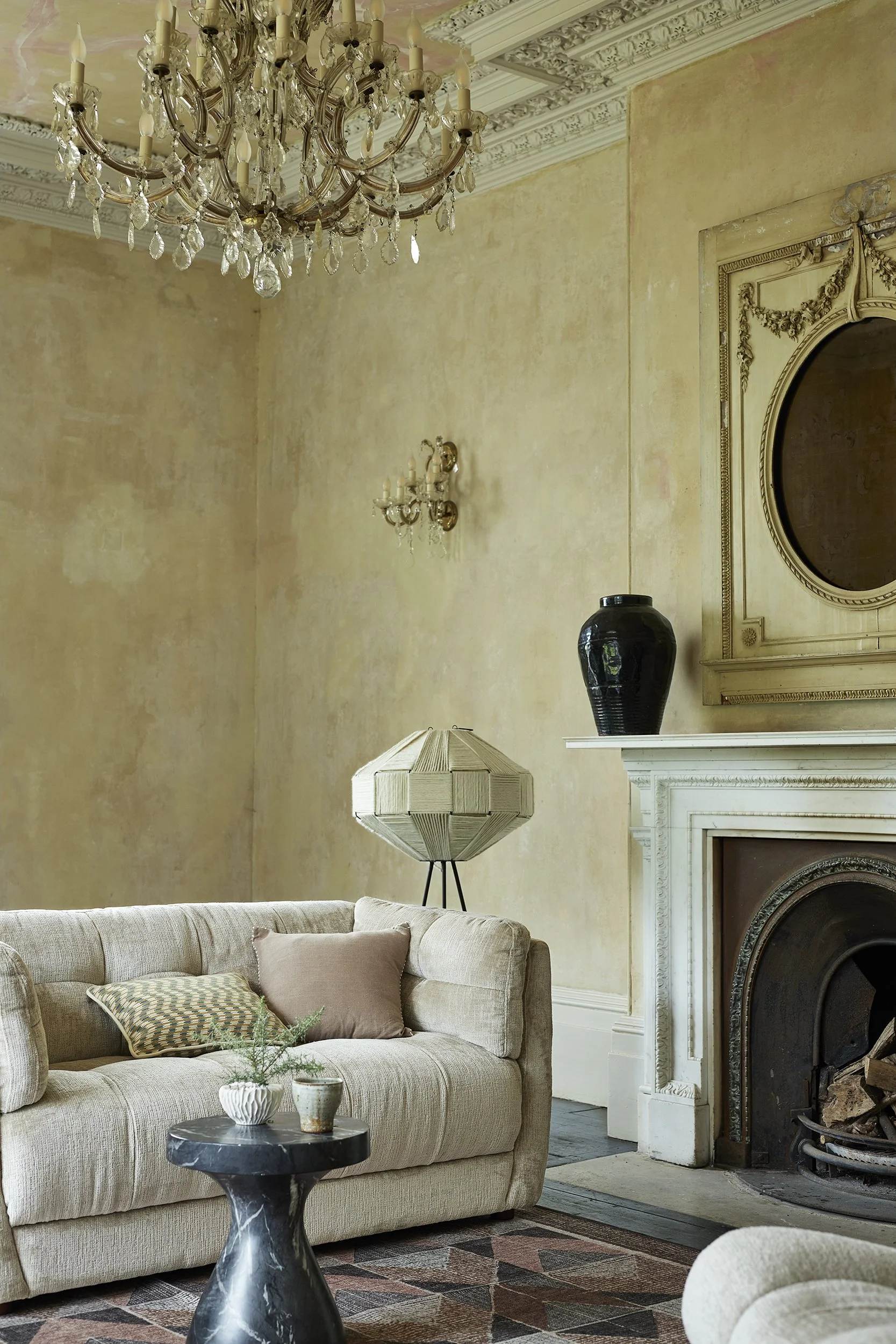 Elegant living room with chandelier, wall sconces, fireplace, beige sofa with pillows, coffee table with plants, vintage decor.