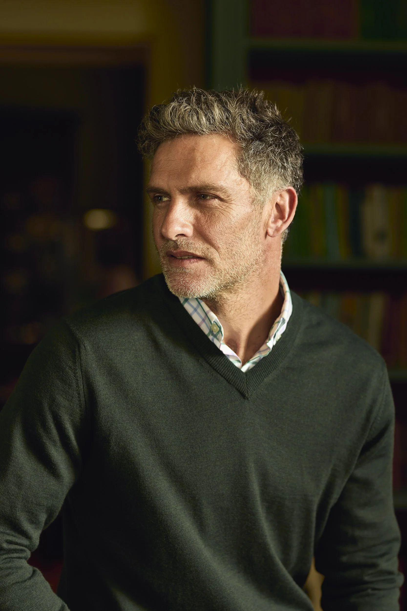 A middle-aged man with gray, curly hair and trimmed beard, wearing a dark green sweater over a collared shirt, standing in a room with a blurred background of bookshelves.