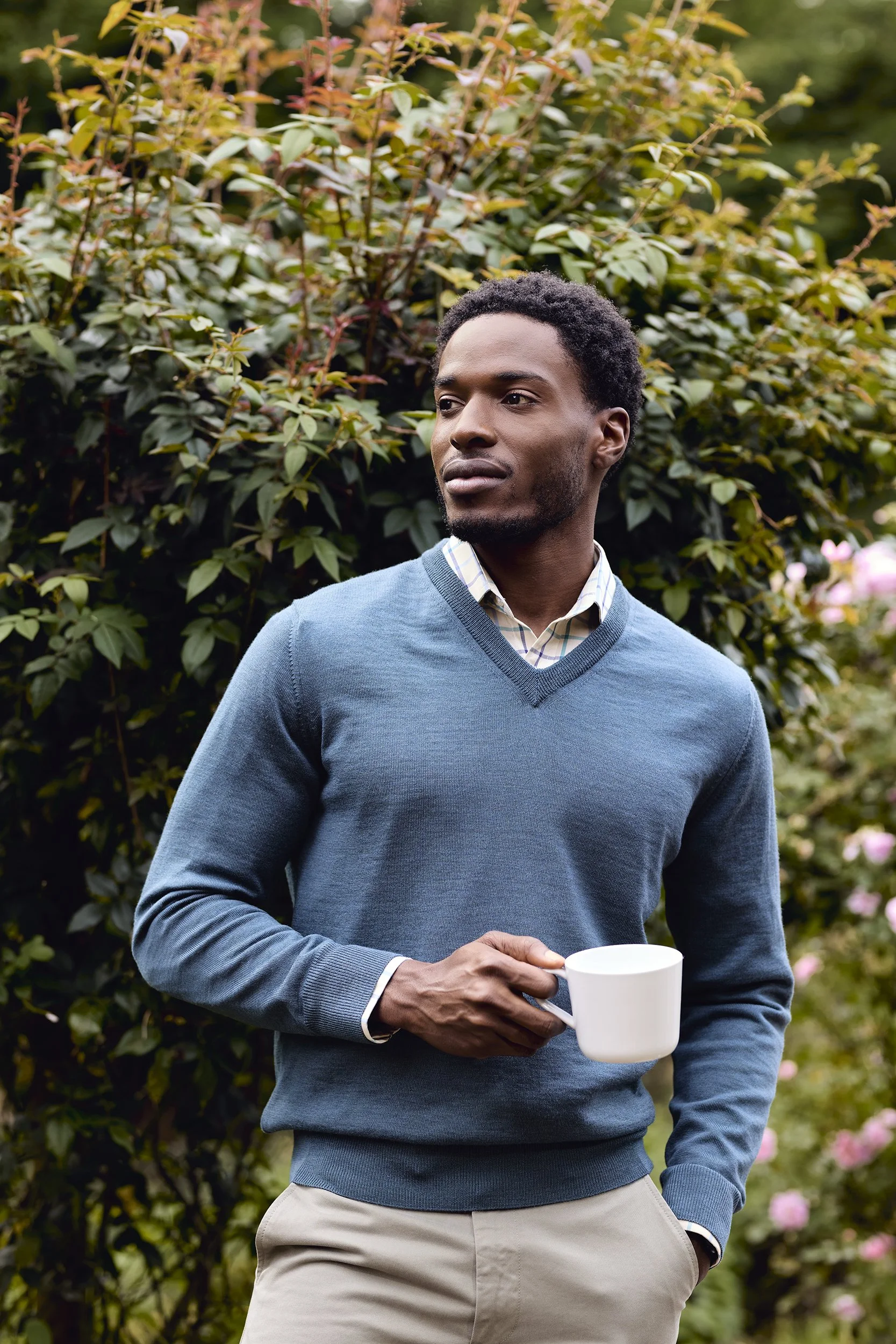 A young man with dark skin and curly hair standing outdoors with greenery and pink flowers in the background, holding a white coffee mug in his right hand, wearing a blue sweater over a collared shirt and beige pants, looking off to the side.