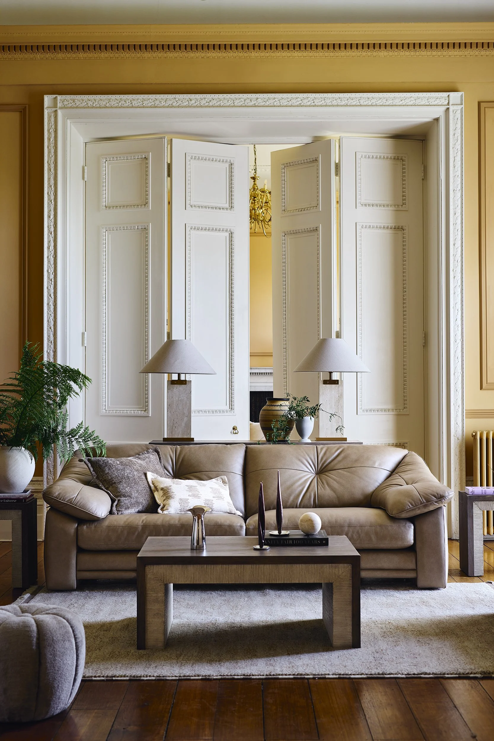 Living room with a beige sofa, decorative pillows, a wooden coffee table with candles and a spherical ornament, side tables with lamps, plants, and open white paneled double doors leading to another room.