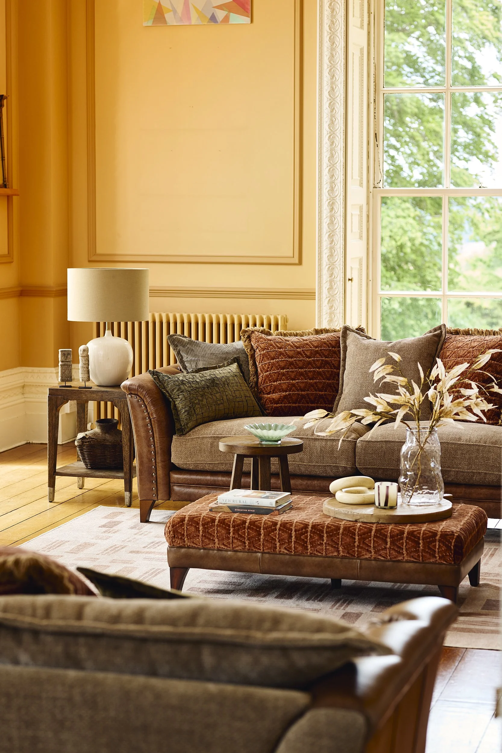 A cozy living room with a tan sofa, decorative pillows, glass vase with dried leaves, wooden coffee table and side table, lamp, window with green trees outside, and artwork on the peach-colored wall.