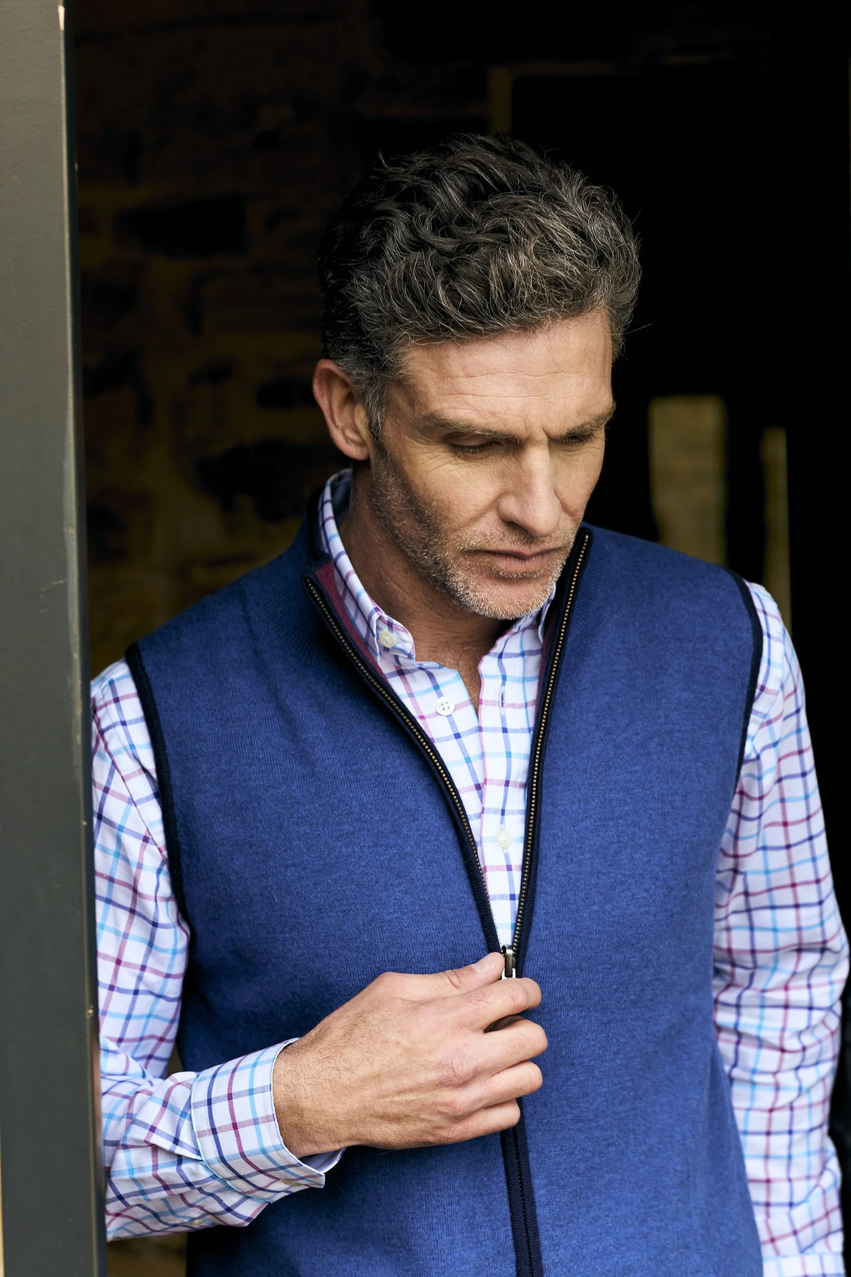 A middle-aged man with salt-and-pepper hair wearing a blue vest over a checkered shirt, looking down and zipping up the vest.
