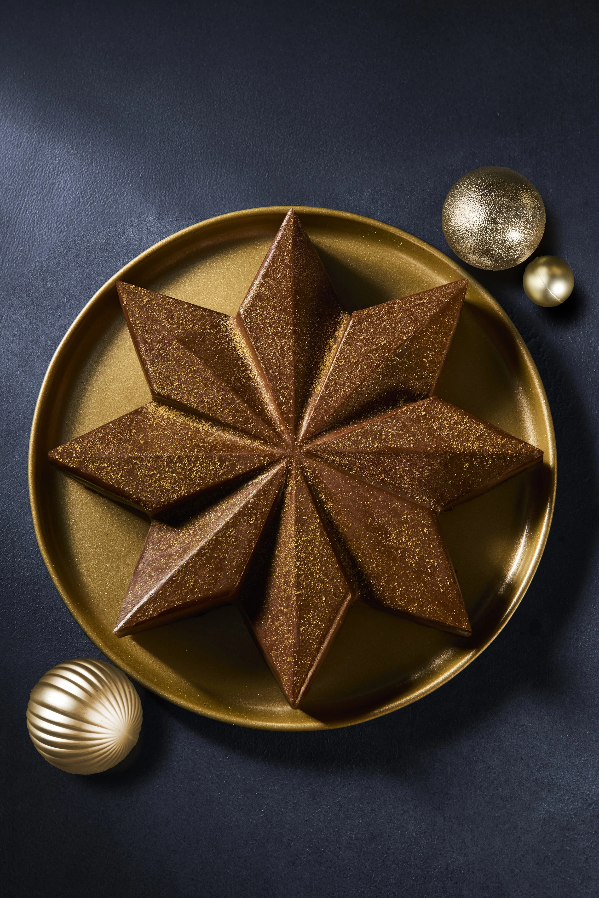 A star-shaped chocolate cake with gold accents on a gold plate, surrounded by gold and silver Christmas ornaments on a dark surface.