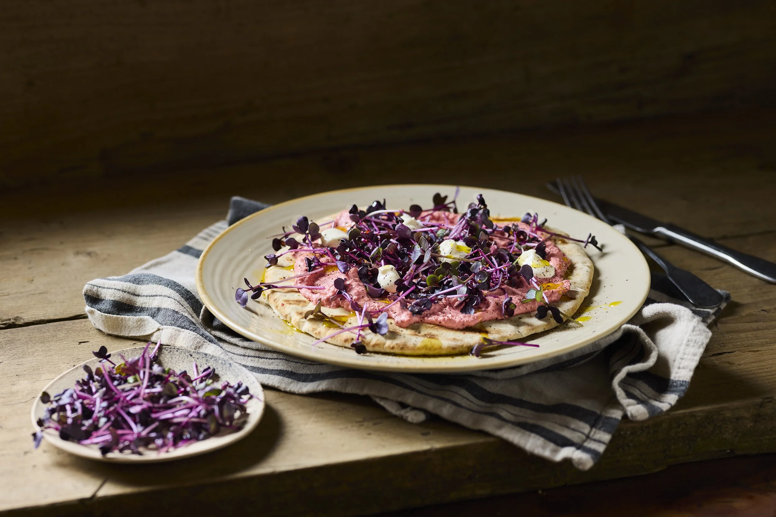 Flatbread pizza topped with a pink creamy sauce, microgreens, and dollops of white creamy cheese, on a yellow plate with a striped cloth underneath, set on a wooden table with a fork and knife